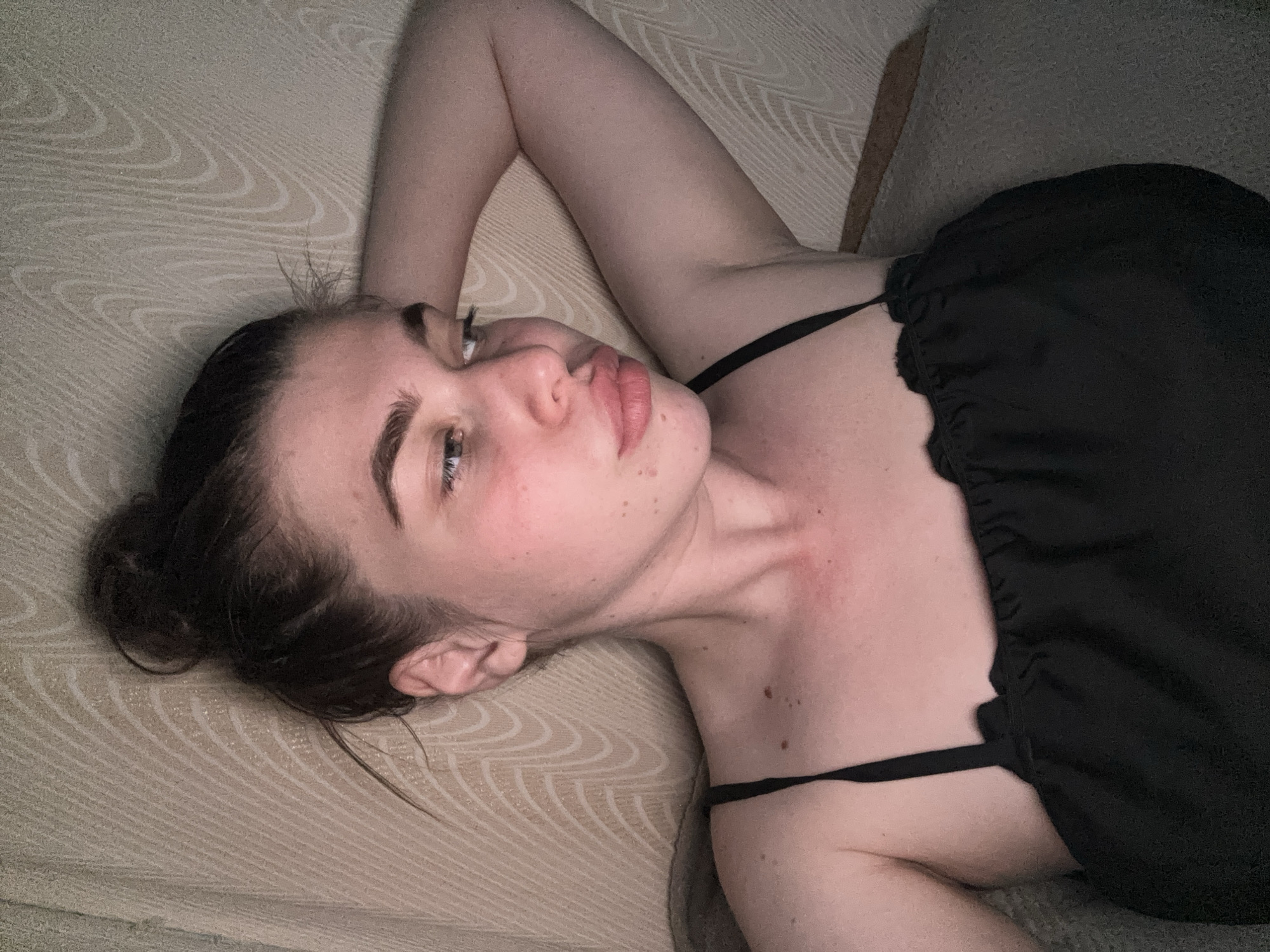 A young woman is lying on a patterned beige surface, possibly a bed or sofa. She is wearing a black garment with thin straps. Her hair is pulled back in a bun. The photograph is taken from an overhead angle. There is no evidence of location in the city of Meshchera, Russia.
