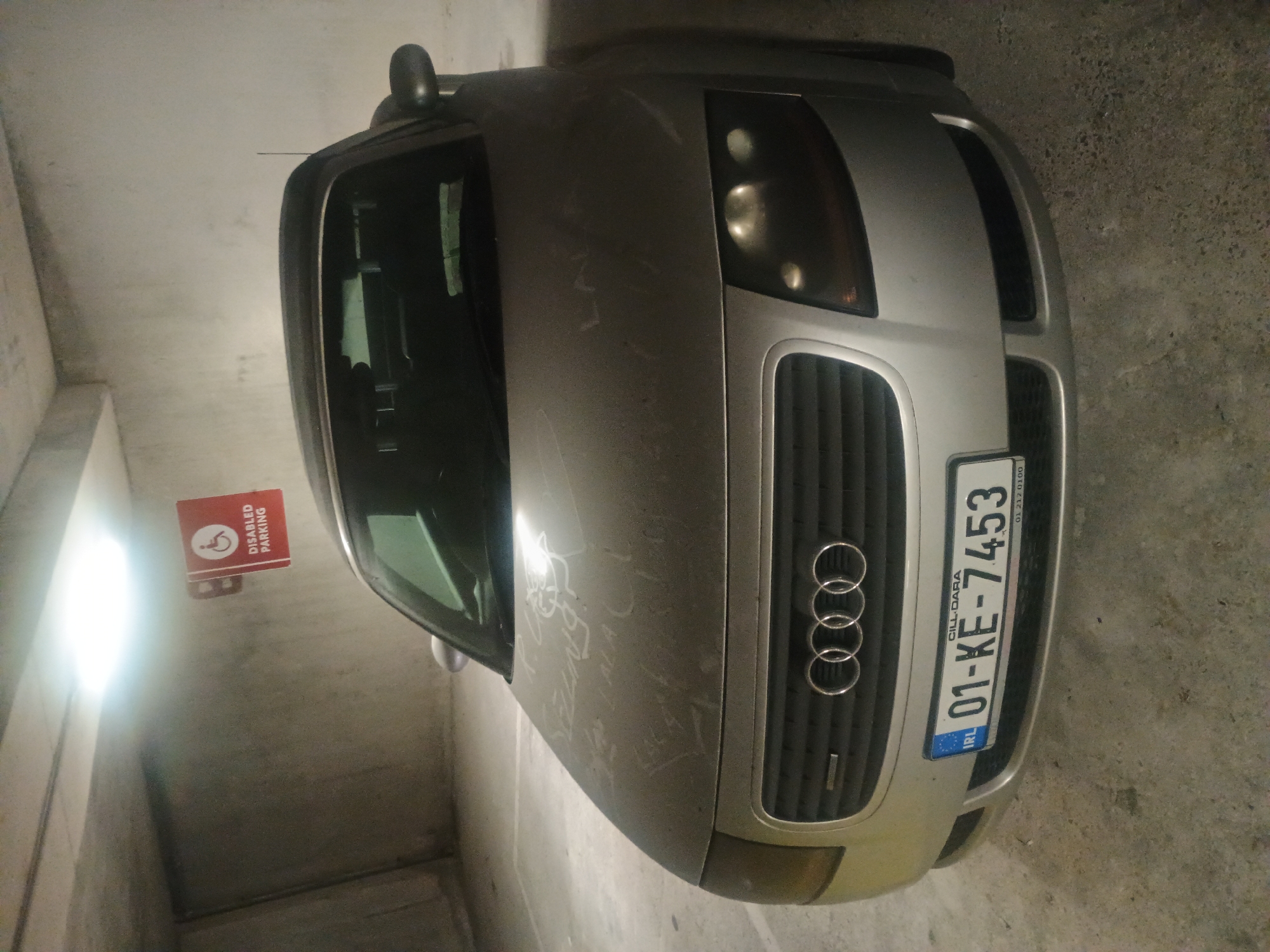 A silver Audi TT convertible is parked in a dimly lit concrete parking garage. The car's hood is covered in dust and has scribbled messages written on it. A red sign indicating "DISABLED PARKING" with a wheelchair symbol is mounted on the concrete wall above and to the left of the car. The car's license plate is visible and reads "01-KE-7453". The floor of the garage is concrete.