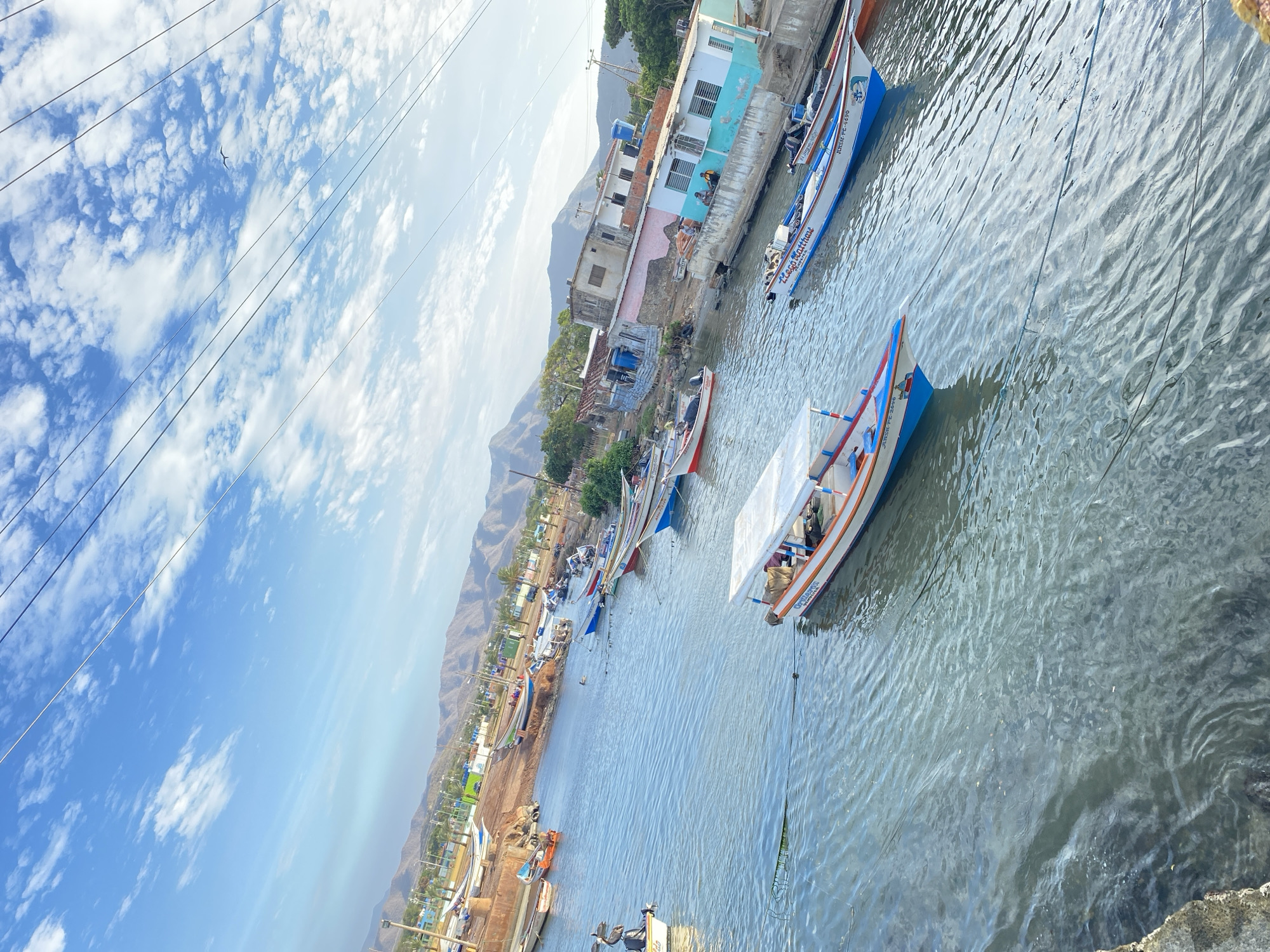The photograph shows a harbor scene in Juan Griego, Venezuela. Several boats, predominantly white with blue and red accents, are moored along a waterway. A prominent boat in the foreground has a white canvas sail and is tethered by a rope. The waterway extends to the shore, where more boats are docked. Buildings, some painted in light blue and white, line the waterfront. In the background, a mountainous landscape is visible under a partly cloudy sky with overhead power lines.