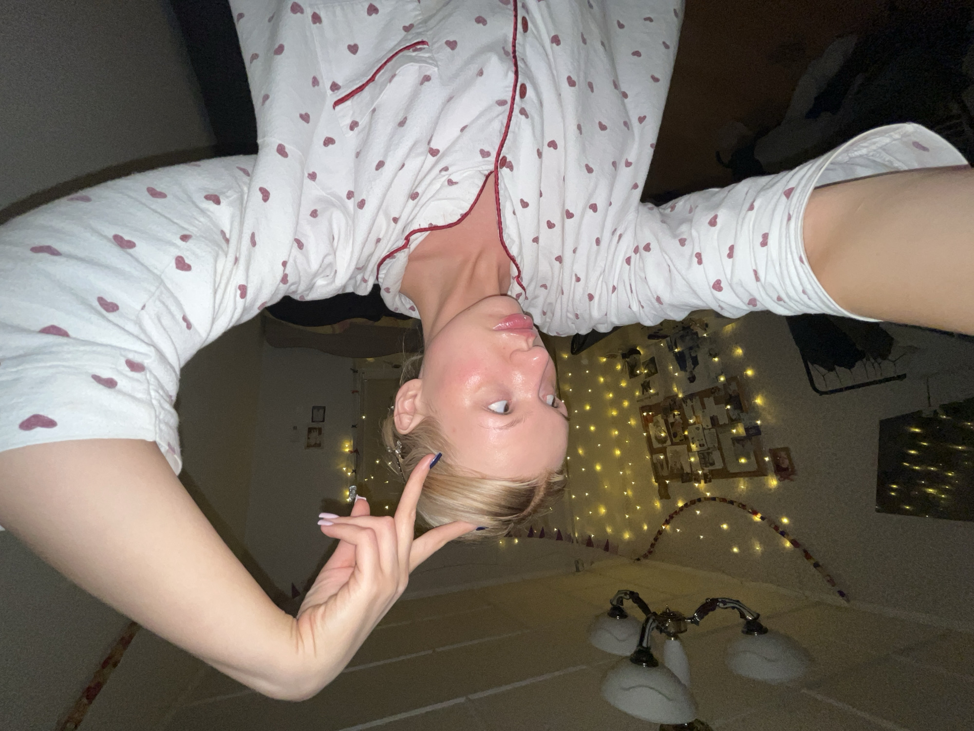 A person wearing a white pajama top with red heart patterns is seen from an unusual perspective, seemingly upside down. The person is indoors; behind them is a bed with a headboard decorated with twinkling string lights. There is a wall with framed pictures and other wall decorations lit by the string lights. A lamp with multiple shades can be seen near the bottom of the frame. The location is potentially a residential setting. The city and location are not ascertainable without further information or image analysis.