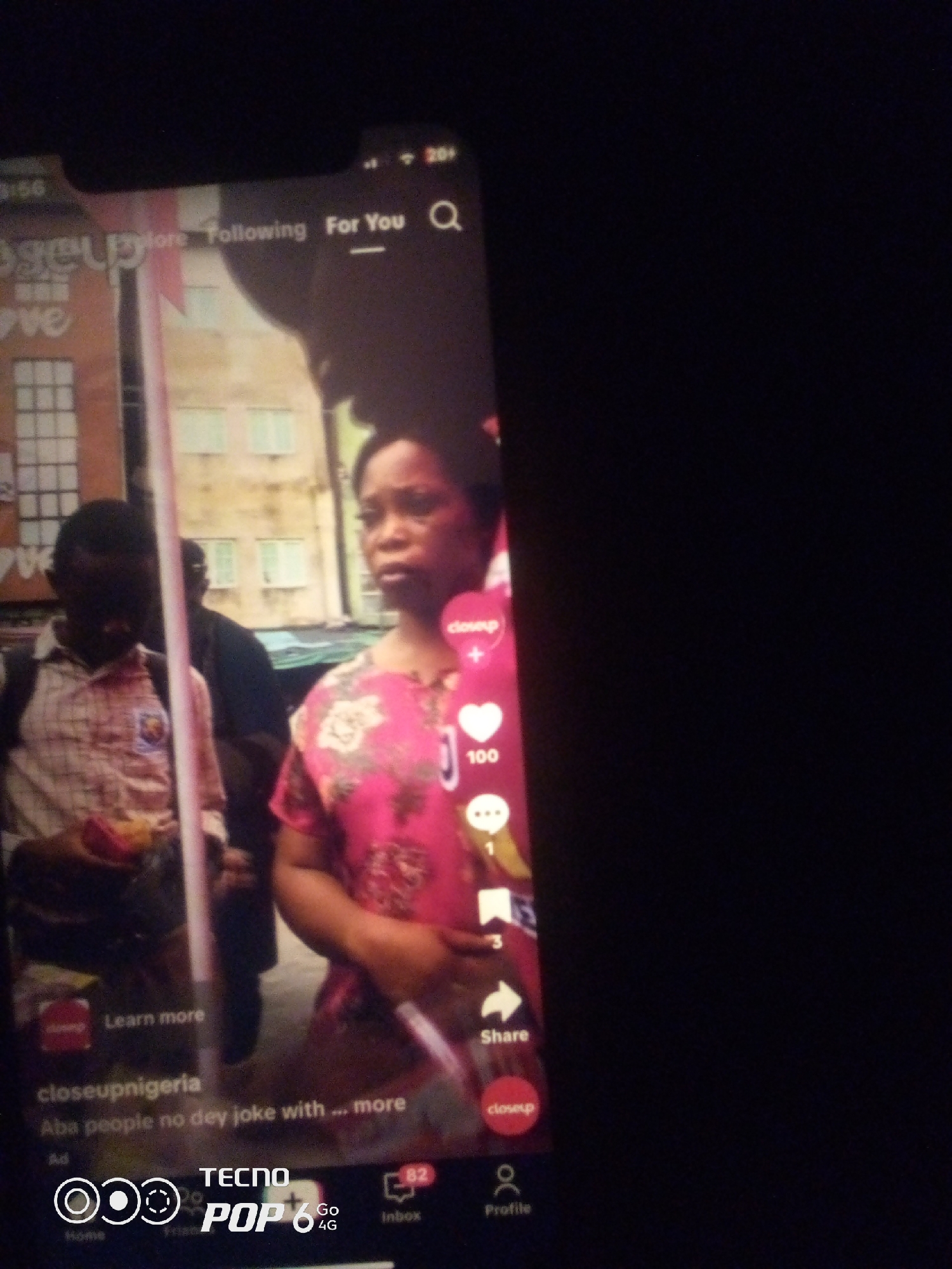The image displays a mobile phone screen showing a social media feed, likely TikTok, with a video playing. A young woman in a pink floral top is the primary subject, looking to the right of the frame. To her left, a young man in a checkered shirt is also visible, holding a small item. In the background, a blurred building with windows can be seen. The interface elements of the app are visible, including icons for likes, comments, shares, and a profile. Text overlays include "Learn more", "closeupnigeria", and "Aba people no dey joke with ... more". The bottom of the screen shows the TECNO POP 6 logo. While the text mentions "Aba people", there is no specific indication of the location being Howayi, Nigeria.