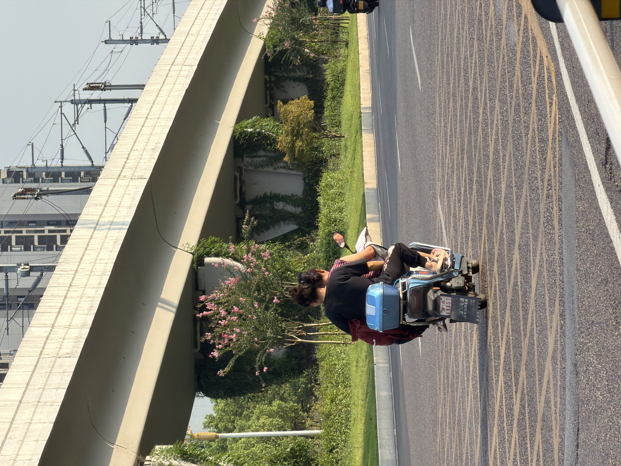 A man and a woman are riding on a scooter on a city street in Jiangjin, China. The man is driving the scooter and the woman is sitting behind him. They are both wearing casual clothes. The scooter has a blue storage box on the back. In the background, there is an elevated highway with buildings and trees. The sky is clear. The street has yellow markings on it, indicating it is a road.