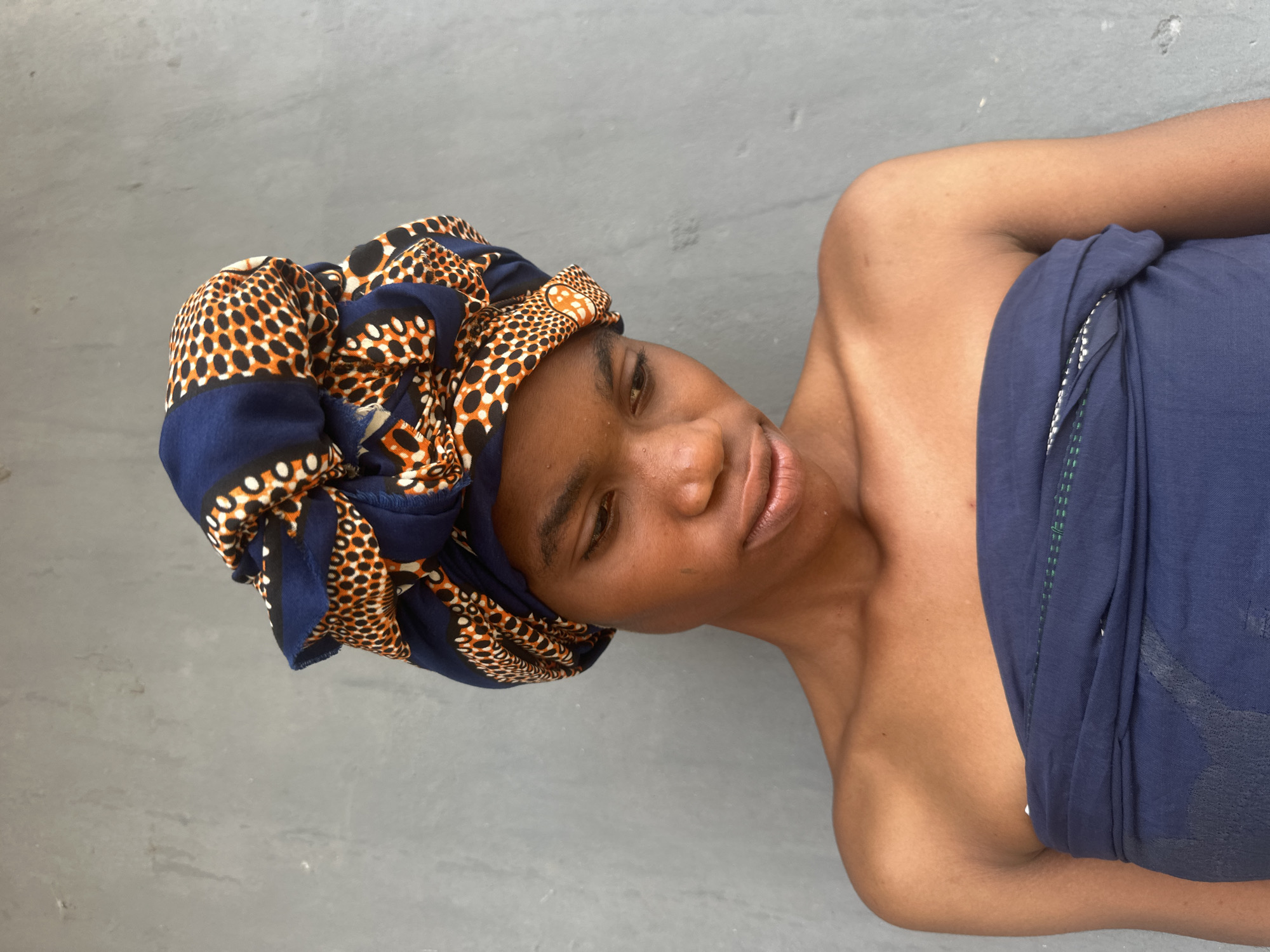 A young person with dark skin, wearing a headwrap with a blue, orange, and white patterned fabric, and a dark blue strapless garment, stands against a grey concrete wall in Badagry, Nigeria. The person is looking downwards with a neutral expression. The lighting suggests an outdoor setting. The image is a medium shot, focusing on the upper body and head of the person.