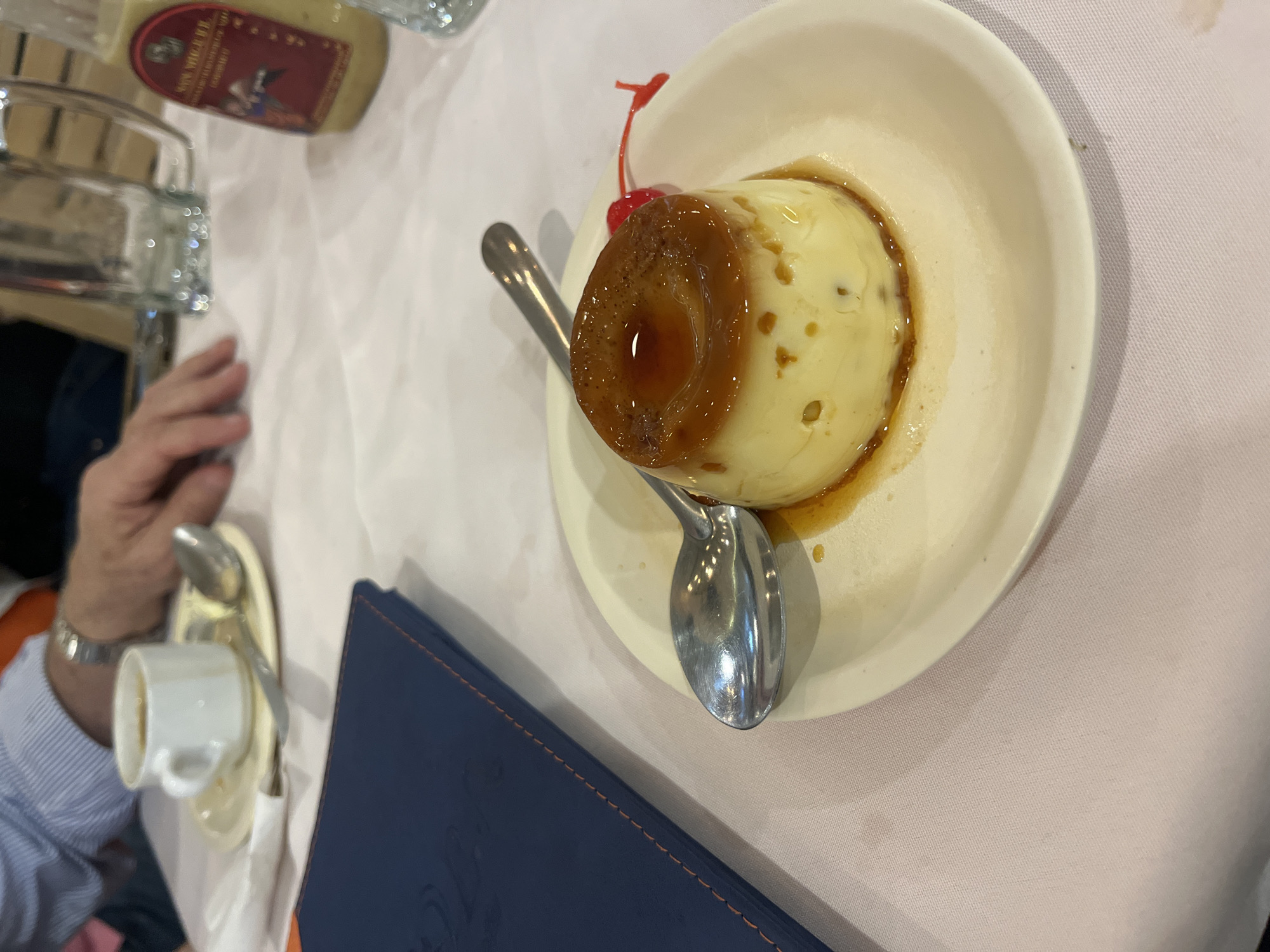 The image depicts a partially eaten flan dessert on a white plate, served with a spoon and garnished with a cherry. A blue menu is situated next to the plate, and there's a small cup of coffee, also with a spoon, nearby. A hand is visible in the lower left corner of the image. In the background, there are bottles and containers on a table covered with a white tablecloth. The image appears to have been taken at a dining establishment. The location is San Bernardino Tlaxcalancingo, Mexico.