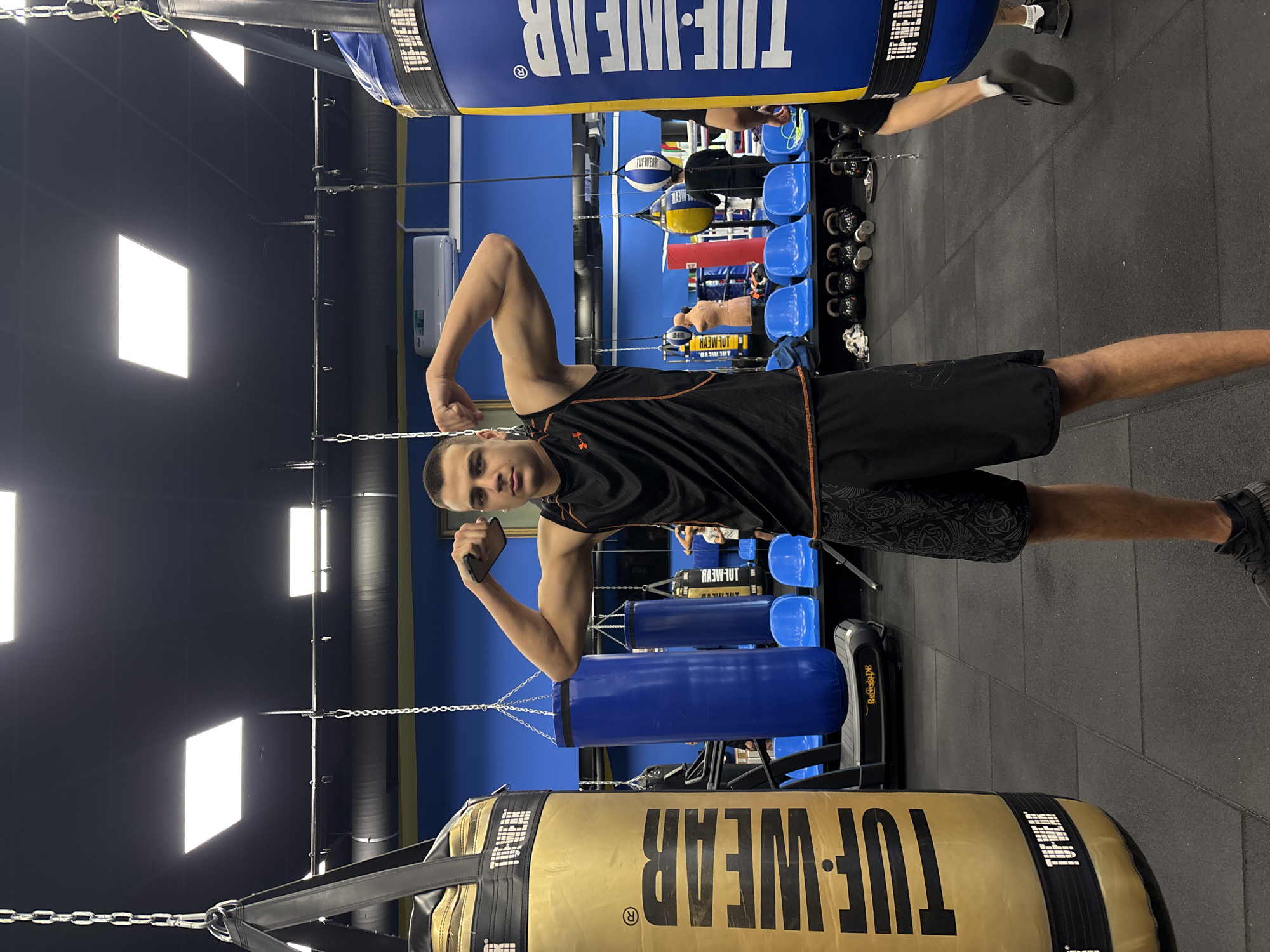 A gym setting in Sofia, Bulgaria, features a man striking a pose between two punching bags suspended from the ceiling. He is wearing a black sleeveless top with orange trim and black shorts patterned with a dragon scale design, and black sneakers are on his feet. His arms are flexed, displaying his biceps. The punching bags, labeled "TUF-WEAR," are different colors, with one being beige and the other blue. In the background, there is an array of blue seating and a rack filled with kettlebells and other gym equipment, positioned on a black tiled floor. The walls are split horizontally, with blue paneling above and black below. Artificial lighting is provided by rectangular panels fixed on the ceiling.