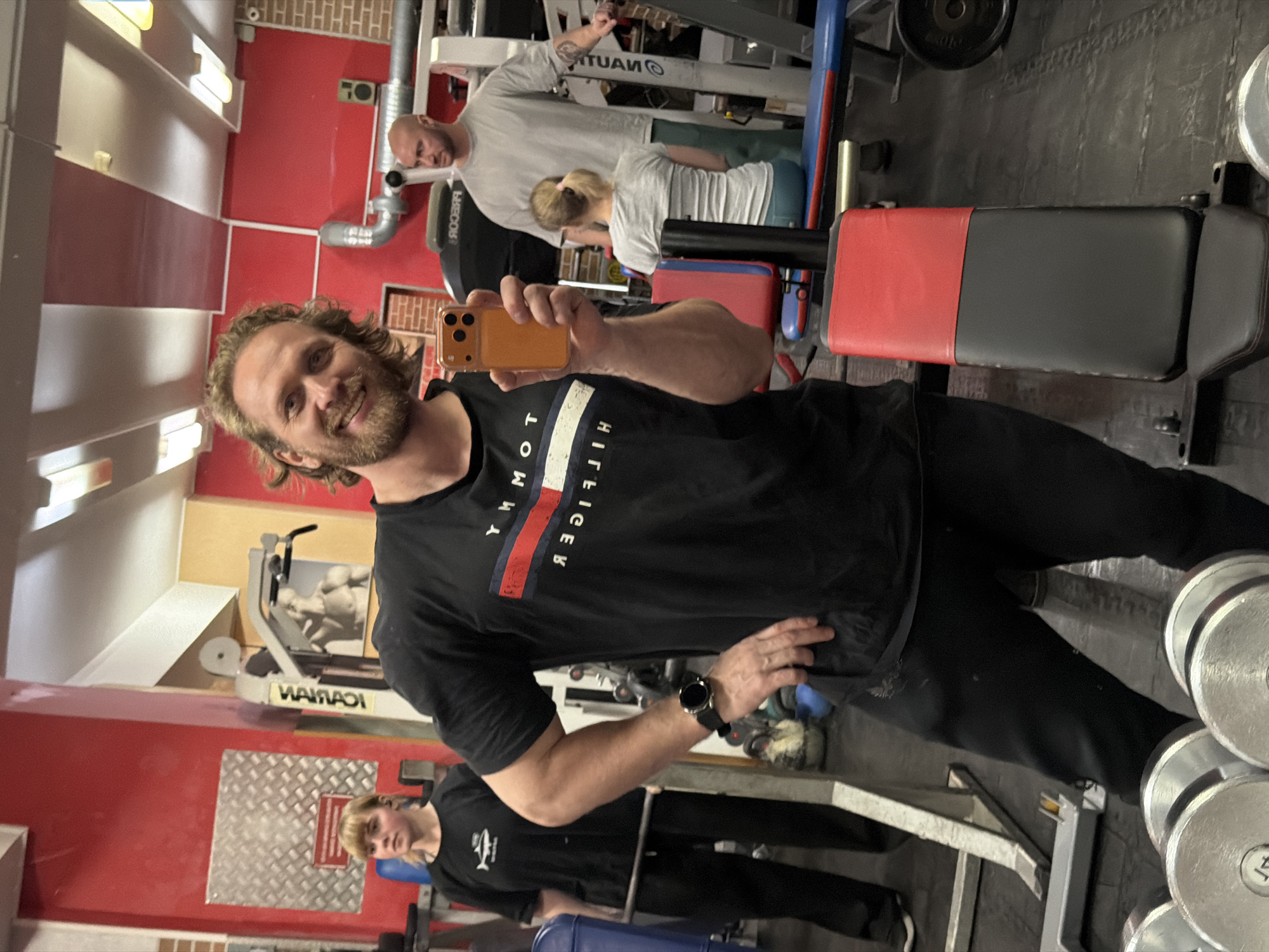 The image is taken indoors in a gym. In the foreground, a man with long hair and a beard is taking a selfie with a yellow smartphone. He's wearing a black t-shirt with a Tommy Hilfiger logo and black pants. On his wrist is a black smartwatch. In the background, another man with a shaved head is working out on a machine. A woman with blonde hair is also present, wearing a white shirt and appears to be working with the man on the machine. A second woman is seen in the background, wearing a black t-shirt. The gym has red walls and visible workout equipment. The city of Saint Petersburg, Russia is not evident from the image.