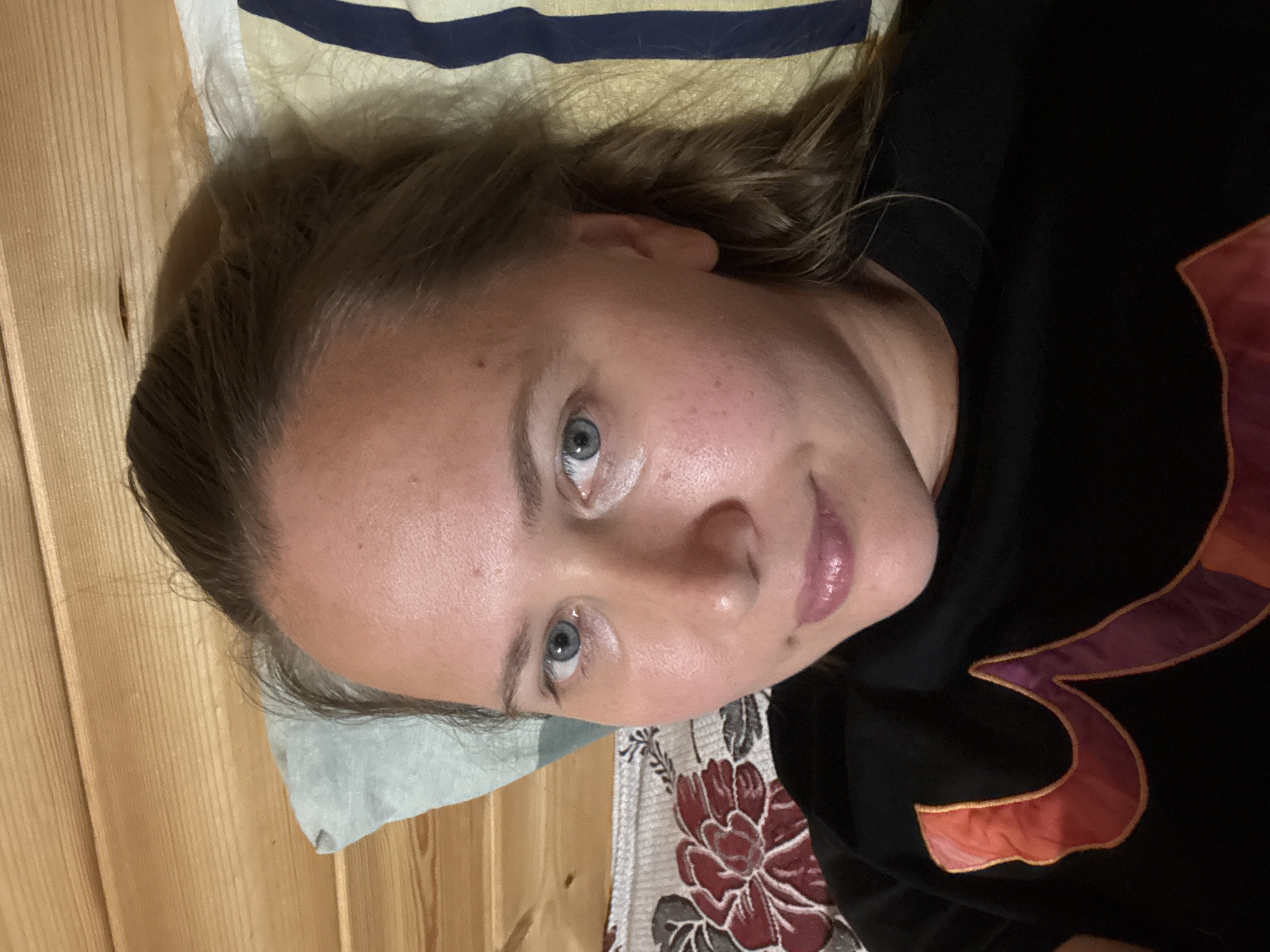 A person is lying down, facing the camera. They have fair skin, light brown hair pulled back, and blue eyes. They are wearing a black shirt with a decorative red and purple design. The person is resting on a pillow with a striped pattern. The background includes wooden paneling and part of a floral patterned fabric. The image does not contain enough information to determine a specific location within Tyulyuk, Russia.