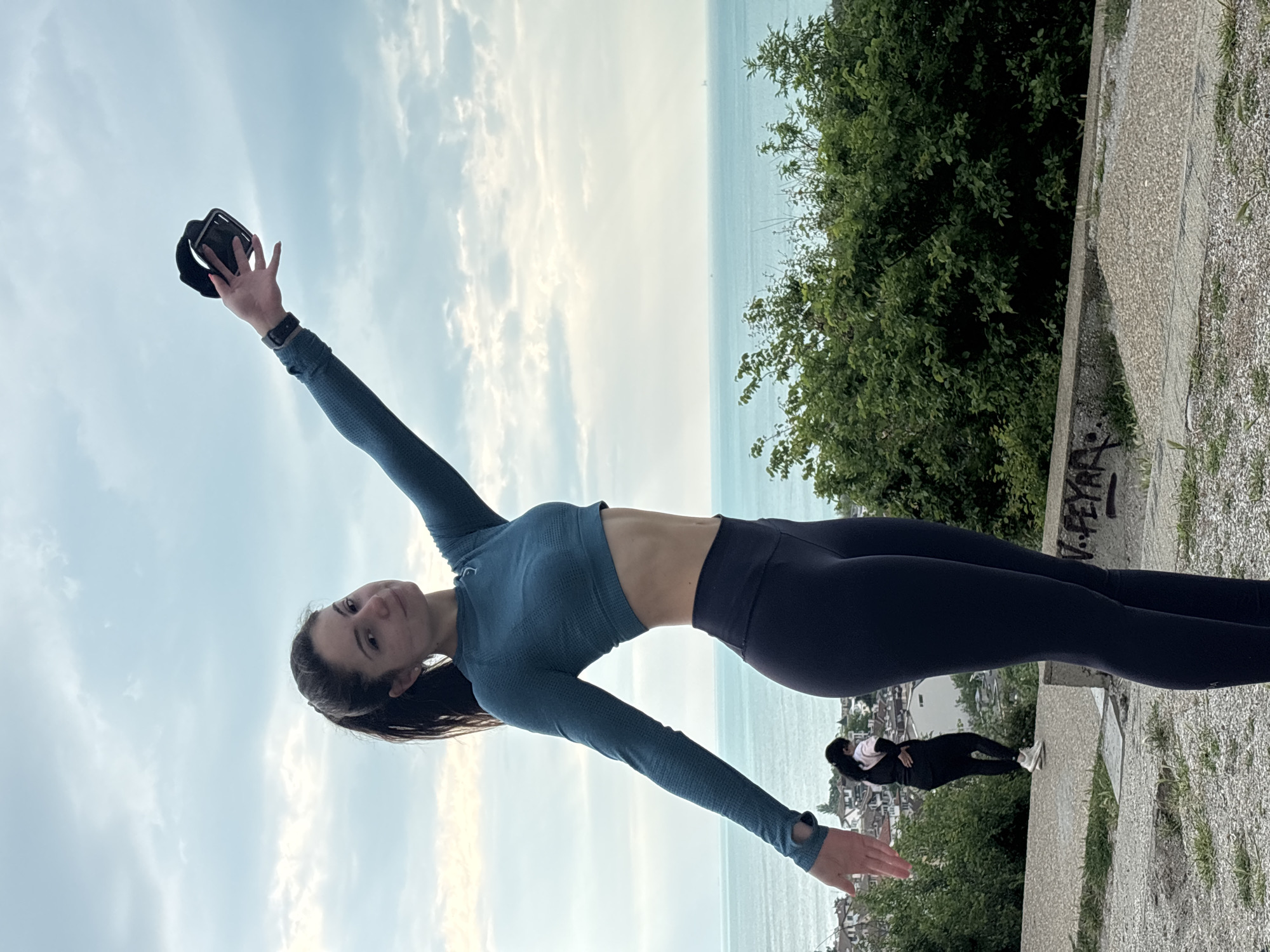 A young woman is standing on a concrete platform or ledge in Varna, Bulgaria, striking a pose with her arms extended. She is wearing a long-sleeved blue athletic top and dark leggings. Her left hand is raised above her head, holding a black object (possibly a hat or sunglasses). The background shows a seascape with a cloudy sky above and a line of green trees and vegetation to the right. Another person is visible in the background walking away.