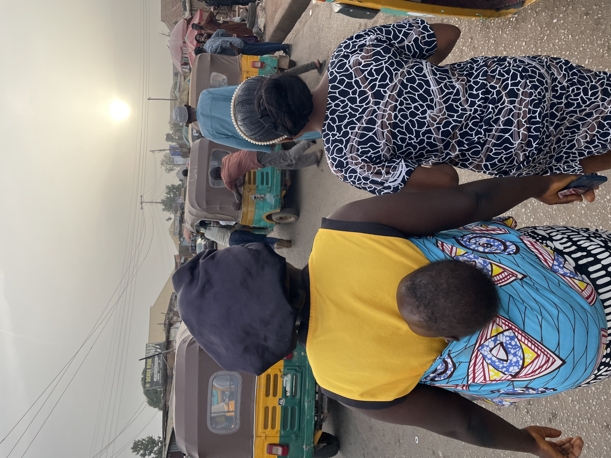 The image depicts a bustling street scene, likely in Jos, Nigeria, captured under a hazy sky. In the foreground, viewed from behind, is a person wearing a bright yellow top who is carrying a baby on their back, secured with a vibrant blue fabric featuring yellow, red, and white geometric patterns. To their right, another individual is seen from behind, dressed in a black and white patterned dress and holding a mobile phone.
Further into the midground, a woman wears a light blue garment and a head covering adorned with pearl-like details. Other pedestrians are scattered throughout the scene, navigating between a row of Keke Napeps (auto rickshaws) which are distinctive to the region. These vehicles appear in various colors like green, yellow, and brown, indicating local transportation.
The background is dominated by a bright, diffused sun in a hazy, light-colored sky, suggesting either early morning, late afternoon, or a day with significant atmospheric dust. Utility poles and overhead wires crisscross the upper portion of the frame. In the distance, buildings and what appear to be market structures are visible, with one sign clearly readable as "DENGE POST." The overall scene conveys the dynamic atmosphere of daily life in a Nigerian urban or market setting.