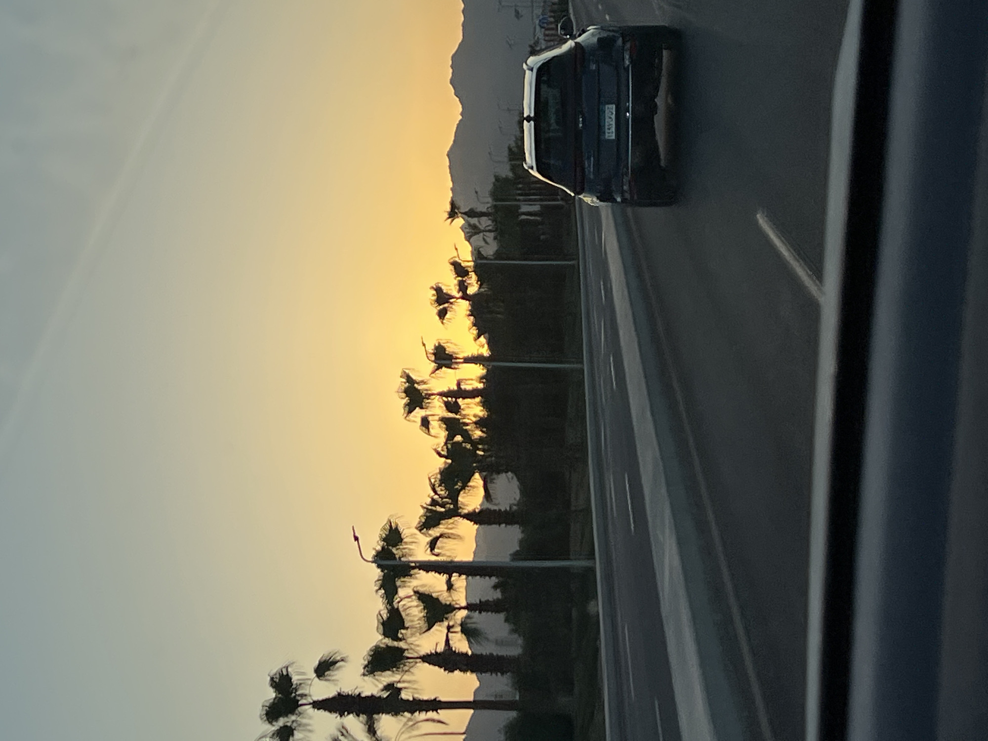 The image captures a scene on a road in Sharm El Sheikh, Egypt, during sunset. A dark-colored SUV is driving away from the camera on the right side of a multi-lane road. Palm trees and streetlights line the left side of the road, with dark, silhouetted mountains visible in the background. The sky is a gradient of pale blue at the top to bright yellow and orange near the horizon, indicating the setting sun. The road itself is paved, with white lane markings visible. The perspective suggests the photo was taken from another vehicle.