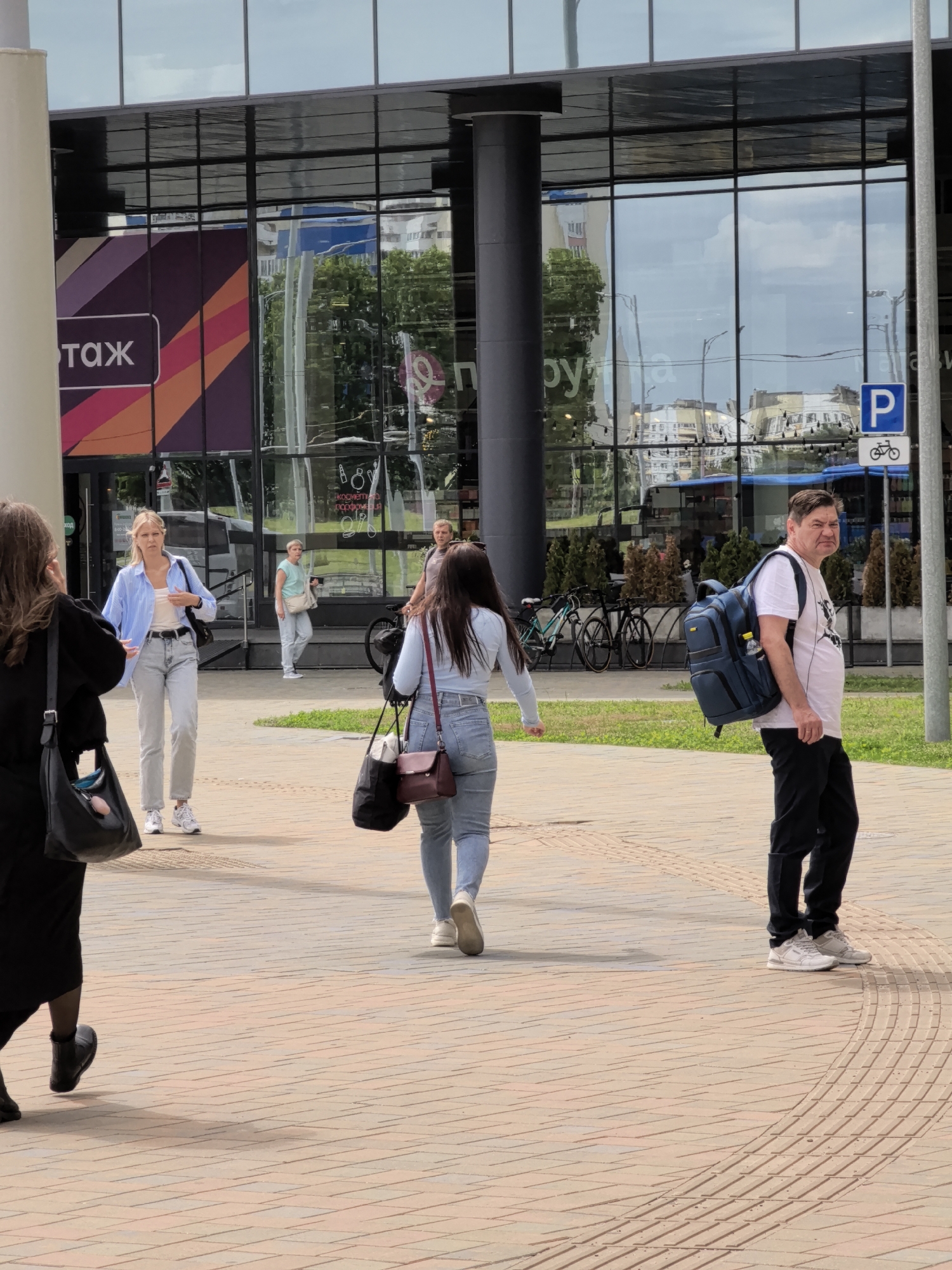 The media file depicts an outdoor scene with several people walking on a paved walkway in front of a modern building with large glass windows. In the foreground, a woman in a black dress walks away from the camera, while a woman in a light blue shirt and grey pants walks towards the camera. Further back, another woman in a light blue shirt and jeans walks away, and a man in a white t-shirt and black pants with a blue backpack walks towards the camera. The building's windows reflect the sky, trees, and surrounding architecture, suggesting it is daytime. There are bicycles parked in front of the building and a parking sign with a bicycle symbol. On the building's facade, text is partially visible, including what appears to be "ОТАЖ" and "Р", suggesting a commercial establishment. The overall atmosphere seems casual, with people going about their day.