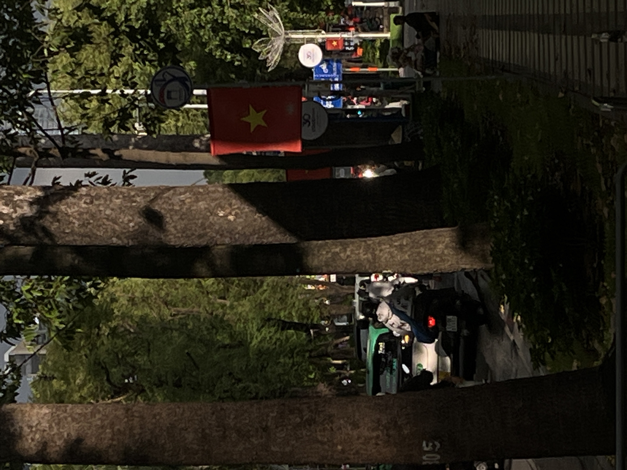 The image depicts a street scene in Thủ Đức City, Vietnam, characterized by a row of large trees lining the roadside. A paved sidewalk runs parallel to the street, with several individuals seated on the ground. A Vietnamese flag is prominently displayed on a pole, accompanied by various street signs. The road itself is active with vehicular traffic, including a green car and several motorbikes, one of which is occupied by two individuals. The overall lighting suggests it is either early morning or late afternoon, with shadows cast by the trees and buildings. Lush green foliage is visible in the background, indicating a well-treed urban environment.