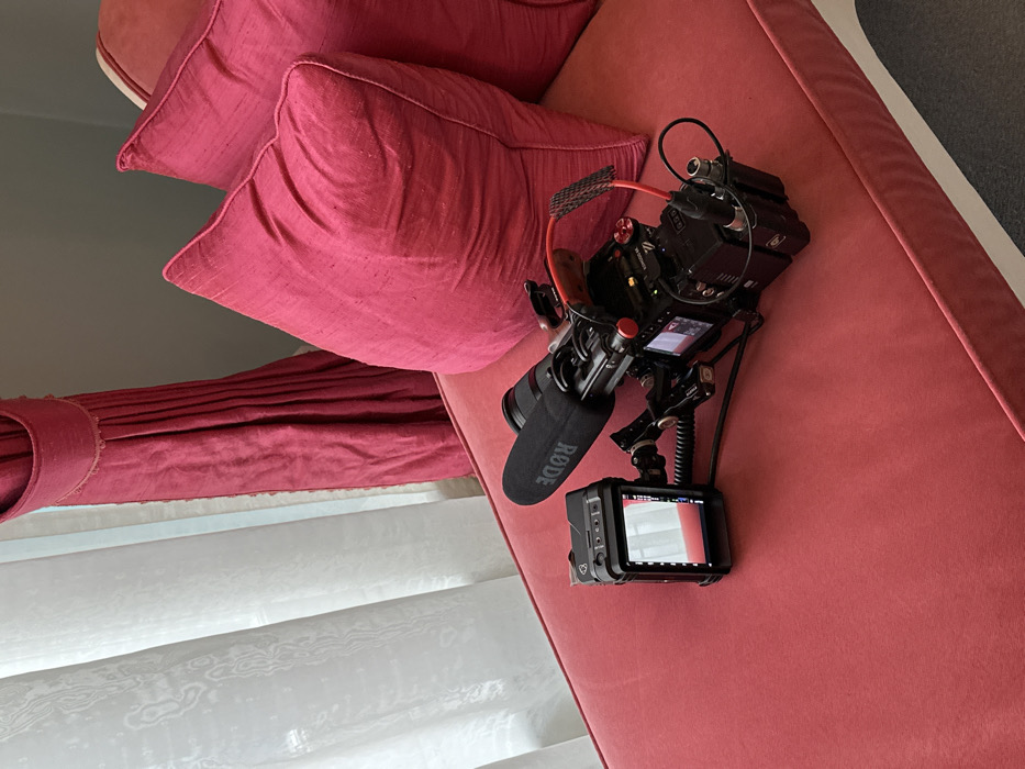 The image displays a professional video camera rig situated on a red upholstered chaise lounge. The rig features a black digital cinema camera body, an attached lens, and a top-mounted Rode VideoMic shotgun microphone. An external square monitor, powered on and displaying a live feed of the surrounding interior, is connected to the camera rig via a coiled cable. Additional camera components, including a cage, a wooden top handle, and a rear-mounted battery pack, are visible. Two matching red throw pillows rest against the back of the chaise lounge. Behind the furniture, a window is covered by white sheer curtains with a textured pattern, while a heavy, pleated red drapery panel is drawn to the right side of the window. The wall visible behind the drapery is a light grey hue. No individuals are present in the scene. This upscale interior setting, indicative of a luxury hotel room, a film production studio, or a private residence, is consistent with environments found within the city of London, United Kingdom.