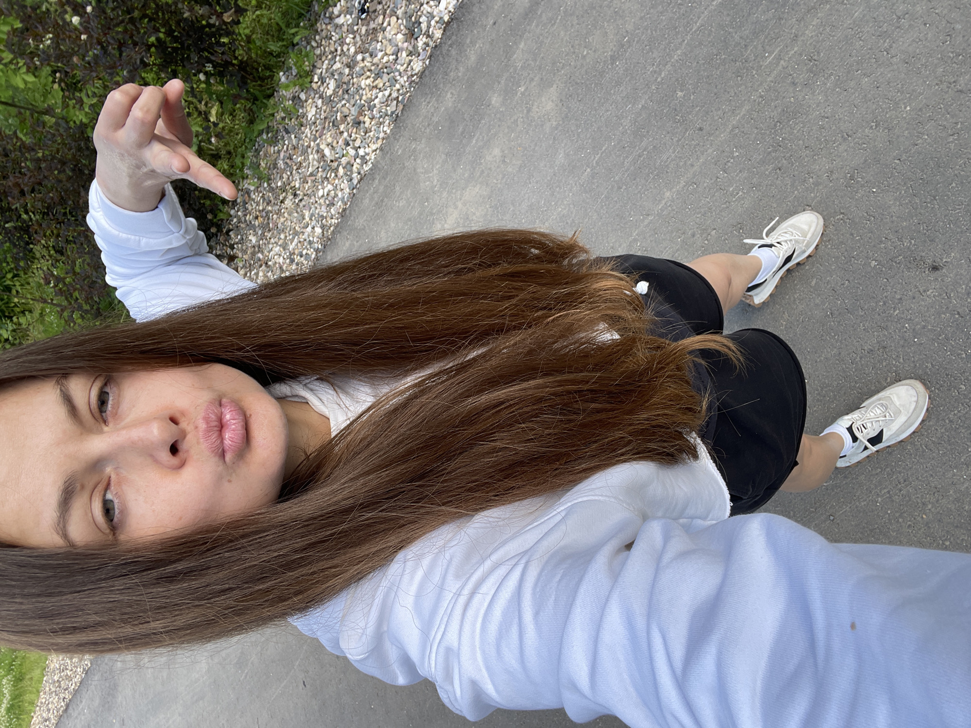 The image shows a woman with long brown hair wearing a white hoodie and black shorts, with white sneakers. She is outdoors, with a paved path and gravelly area in the background, suggesting a park or a pathway. The woman is posing with her lips puckered and one hand held up in a gesture. She is on a paved walkway. There are bushes with dark leaves and some small white flowers visible in the background to the right.