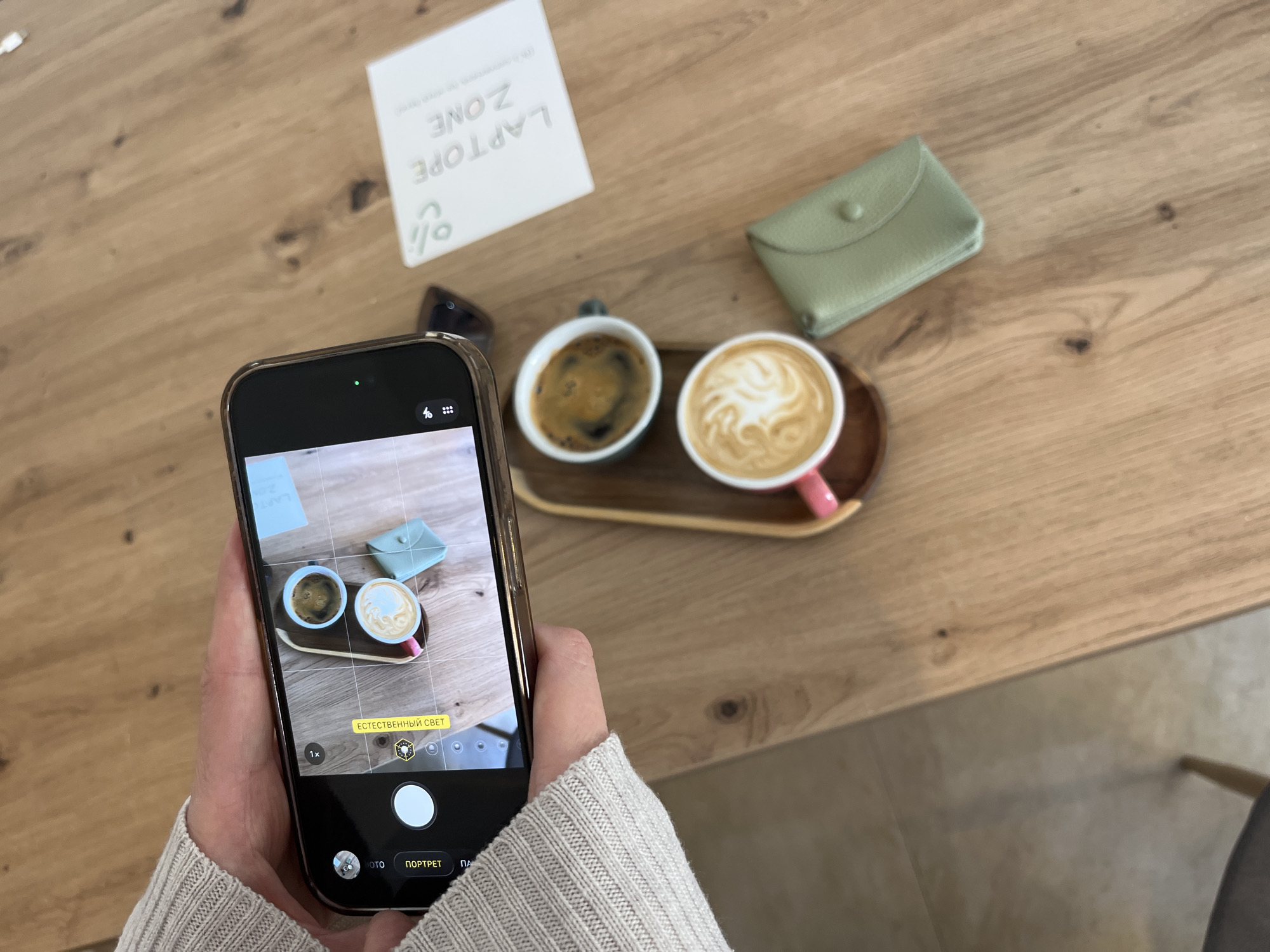 A person in a beige sweater is using a smartphone to take a photo of two cups of coffee on a wooden tray. Next to the coffee, on the wooden table, is a small, light green wallet and a white card that reads "Laptop Zone." The coffee cups appear to contain espresso and latte. The environment is a café with a light wooden table, likely located in Budva, Montenegro.