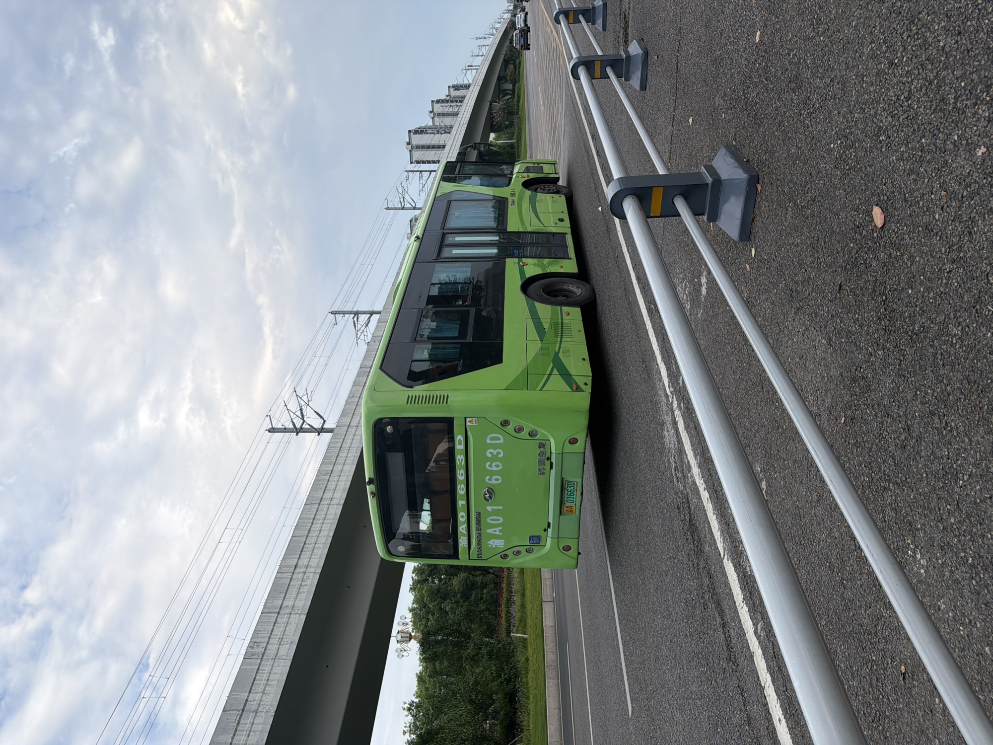A green public bus is traveling on a road in the city of Jiangjin, China. The bus has the license plate "A01 663D". It is a sunny day with clouds in the sky. The road is lined with guardrails and there are trees and buildings in the background. An overpass is visible in the upper left corner of the image, with power lines running above it.