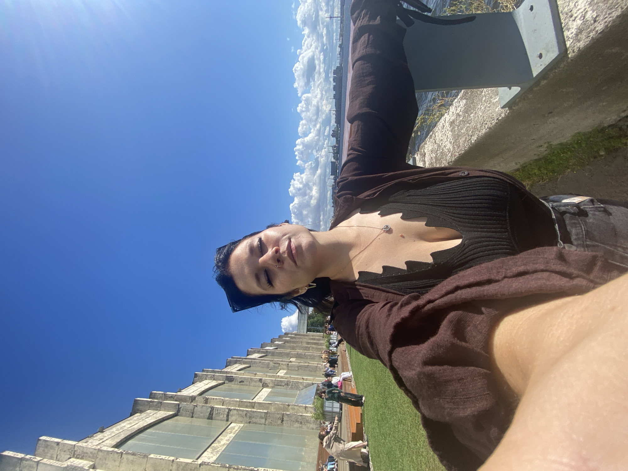 The image captures a selfie of a woman outdoors on a sunny day in St. Petersburg, Russia. The woman is wearing a dark, low-cut top and a brown cardigan. She has dark hair and is wearing sunglasses on her head. She is standing near a concrete barrier overlooking a body of water.
In the background, there is a large, light-colored industrial building with many windows. A grassy area with people seated on wooden steps is visible in front of the building. The sky is a clear blue with scattered white clouds, and the sun is shining, casting a bright glare on the right side of the image. The overall scene suggests a public outdoor space, possibly a waterfront park or recreational area adjacent to an industrial complex. The city skyline is visible in the distance across the water.