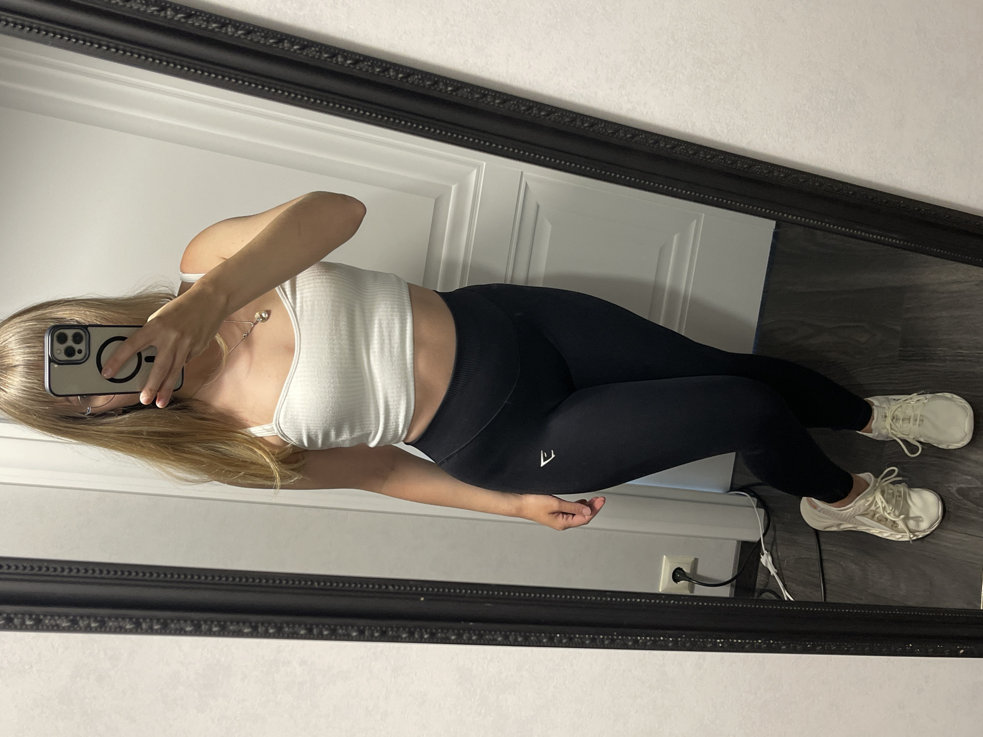 A young woman with blonde hair, wearing a white crop top and black leggings, is standing and taking a mirror selfie. She is wearing white athletic shoes. The background appears to be an indoor setting, possibly a room in an apartment or house, with a white wall and a dark wooden floor. A large, ornate black mirror is positioned to the left of the frame, reflecting the woman and the surrounding environment. A white electrical outlet with a black plug and white wires is visible on the wall near the floor. The scene does not provide any specific visual cues to determine its location within the city of Zhytomyr, Ukraine.