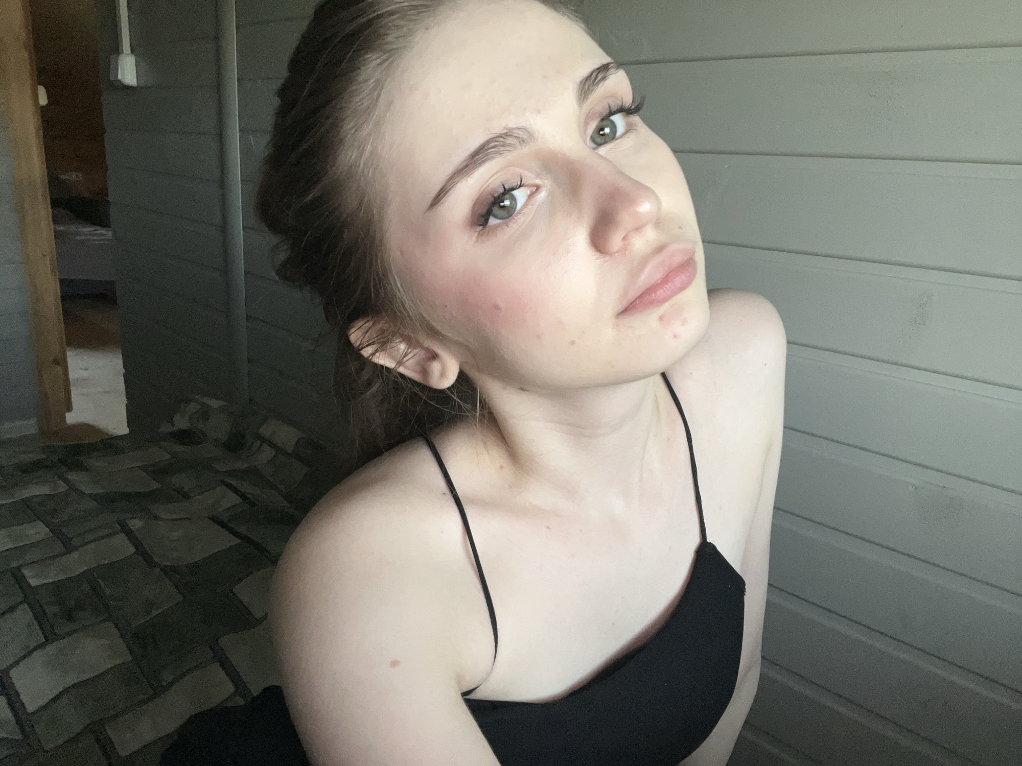 This media file is a close-up, high-angle, selfie-style portrait of a young woman. She has light skin, light brown hair pulled back, and light gray-blue eyes. Her makeup includes mascara, eyeliner, and blush. She is wearing a black camisole with thin spaghetti straps. The background consists of a grey, horizontal-slatted wall and a patterned floor that appears to be tiled or made of wood. To the left, a wooden doorway or frame is visible, leading into a dimly lit interior with what looks like a bed. The lighting suggests it is daytime, possibly indoors or in a shaded outdoor area. The woman's expression is neutral to slightly pouty, and her gaze is directed towards the camera. The scene is likely in Ashkadar, Russia. There is no visible text.