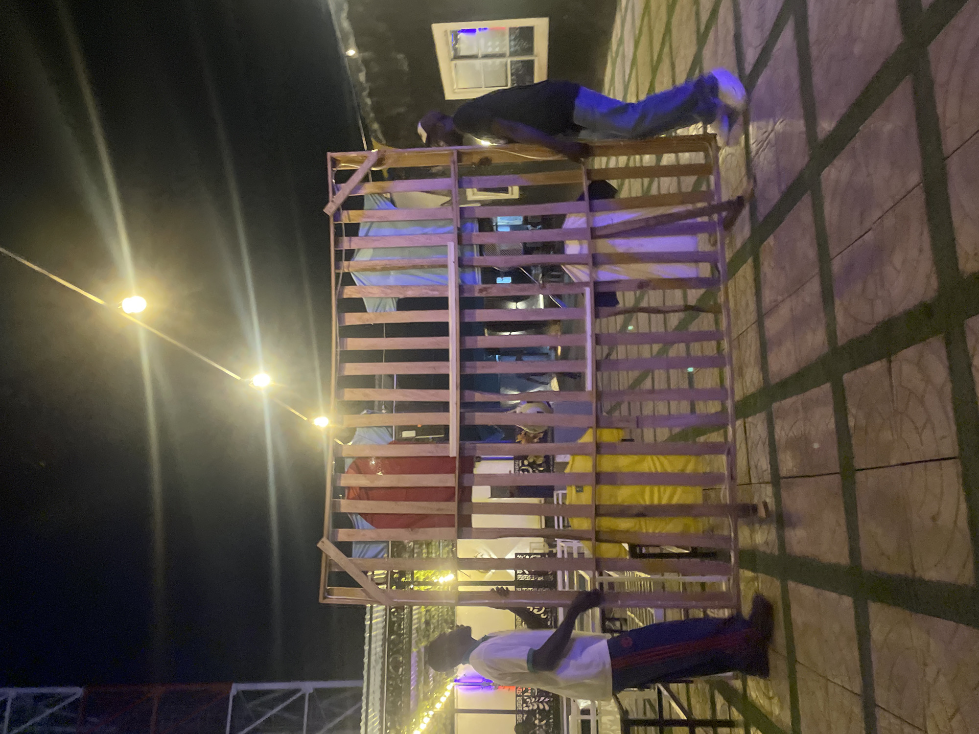 The image depicts an outdoor scene in what appears to be Bar Jirgi Summa, Nigeria, given the context. Two individuals are interacting with a large, multi-tiered wooden structure. One person, wearing a dark shirt and jeans, is positioned on top of the structure, appearing to be leaning over the edge. The second individual, wearing a white shirt and patterned pants, is standing at the base of the structure, looking upwards. The wooden structure is composed of multiple horizontal wooden slats arranged in a grid-like pattern. Colorful fabrics or materials are visible within the structure's framework. In the background, lights strung overhead illuminate the scene, and part of a building with a window is visible. The ground is paved with patterned tiles. A glimpse of additional structures is seen in the lower portion of the image. The image appears to be taken at night or in low-light conditions.