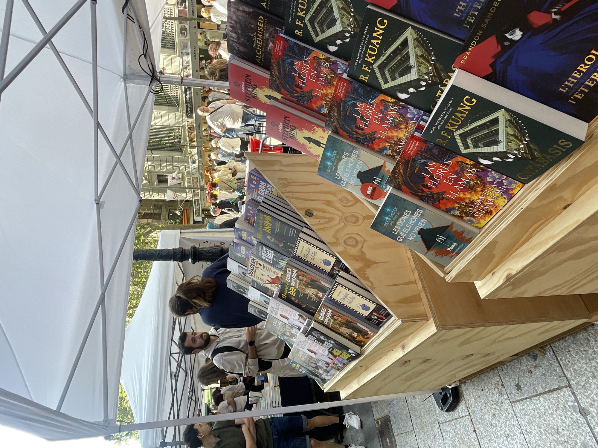 This medium shot depicts an outdoor book market stall, likely part of a "Feria de Artesanos" (Artisan Fair) in Barcelona, Spain. The scene is bustling with people browsing a wide selection of books displayed on wooden shelves under a white pop-up tent.
In the foreground, a prominent wooden display holds numerous books, many of which are oriented to show their covers and spines. Visible titles include "Las Flores en Llamas" by Samantha Shannon, multiple works by R.F. Kuang (including "Catadas," with a banner "AUTORA SUPERVENTAS NÚMERO 1 DEL NEW YORK TIMES"), "Alchemised," and "Carrie" by Stephen King. Other authors like Brandon Sanderson ("L'Heroi L'Etern," "Mistborn") and Alice Sheldon ("Les Dones Que Els Homes No Veuen") are also represented. The books are predominantly in Spanish or Catalan. Cookbooks such as "52 Cuines Pim Pam" and "Els Plats del Dia" are also displayed.
Several individuals are engaged in browsing the books. A man with a beard and a light-colored top leans over the display, while another person with long, dark hair also examines the selection. In the background, more market-goers can be seen under additional white tents, suggesting a lively outdoor event. Architectural elements like buildings with windows and balconies line the street beyond the tents.
The scene is bathed in bright daylight, indicating clear weather. The ground is paved with grey tiles. Visible signs in the background read "Feria de Artesanos," "TARLATANA," and "COMABARC," providing further context to the market setting. The overall atmosphere appears calm, with people focused on discovering new books.