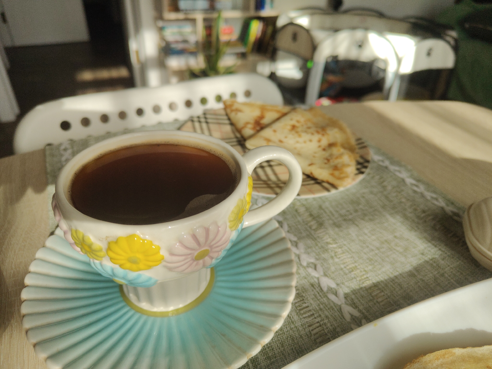 The image captures a close-up view of a coffee cup and saucer on a table. The cup is white with colorful floral decorations, and it is filled with dark coffee. The saucer is a pale blue with a ribbed design. Behind the cup and saucer, on a checkered placemat, are two folded crepes. The table appears to be made of wood and is bathed in sunlight, casting shadows. In the background, out of focus, are hints of indoor furniture, possibly chairs and shelves with books, suggesting a domestic setting. The scene does not provide any information to place it specifically in the city of Yuzhny, Russia or any other specific location.