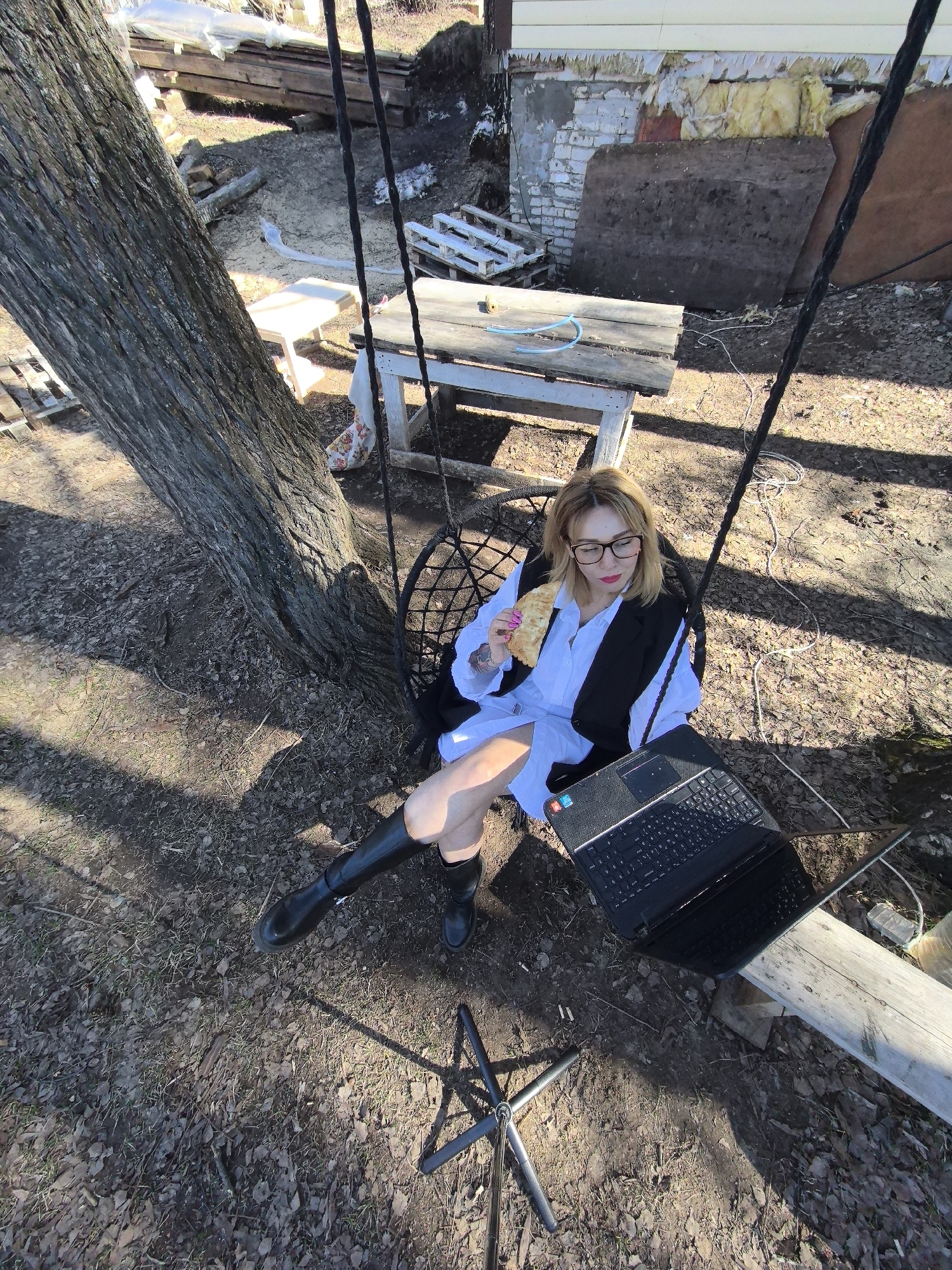 A woman sits in a black, hanging chair in an outdoor setting, with a large tree to her left. She is fair-skinned, wearing glasses, a white shirt, a black vest, and black boots, and is holding a pastry in her left hand. A black laptop rests on her lap. The ground is covered in fallen leaves and twigs. In the background, there's a building foundation with exposed insulation, wooden planks, and various pieces of debris. The location appears to be a backyard or wooded area, which does not provide enough information to determine if the scene is in Noviy, Russia.