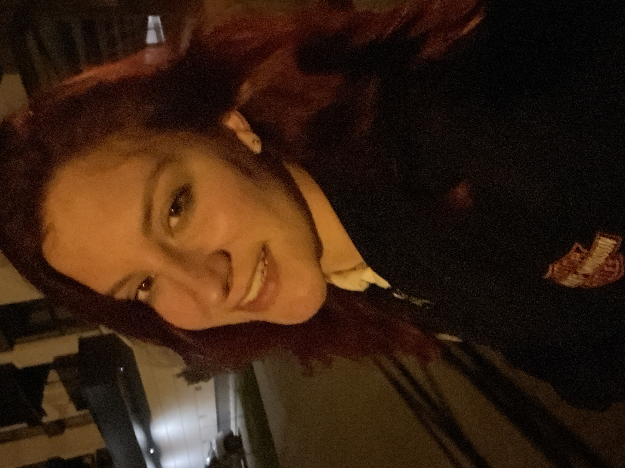 A woman with auburn hair smiles at the camera, wearing a dark jacket with a Harley Davidson logo patch visible on the right side. The background suggests an outdoor setting, possibly at night, with indistinct urban elements. Details about the specific location in Asenovgrad, Bulgaria, are unavailable from the image.