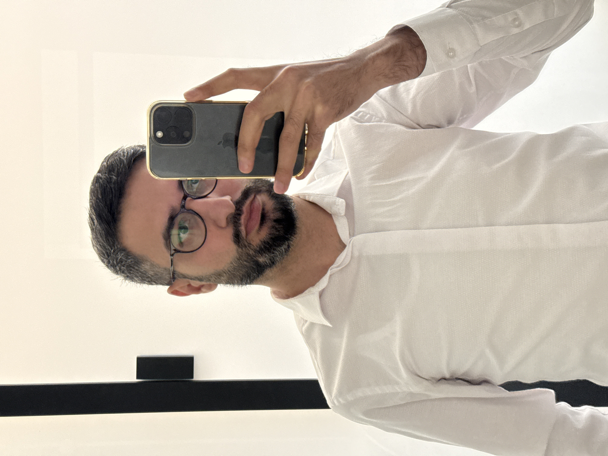 The image captures a mirror selfie of a man, oriented sideways due to the reflection angle, in an indoor setting in Milan, Italy. The subject is a man with short, dark hair showing some grey, a well-groomed dark beard, and round-rimmed glasses. His eyes, visible through the lenses, appear focused, and his lips are slightly parted. He is wearing a crisp white long-sleeved collared shirt.
His right hand holds a dark-colored smartphone, likely an iPhone given the distinctive camera module and the subtle outline of the Apple logo visible on its back. The phone is encased in a gold or light-colored protective cover. The man holds the phone up, seemingly pointing it towards his reflection to capture the image.
The background is predominantly a bright, light-colored wall or surface, suggesting a well-lit environment, possibly with ample natural light during the daytime. Below the man's head and shoulder, a dark, rectangular object, possibly a shelf or part of a furniture piece, runs horizontally across the frame, with a smaller dark block sitting on top of it. The overall impression is a casual, well-lit portrait taken in an interior space. No discernible weather conditions are visible, and the emotion conveyed is neutral or slightly inquisitive.