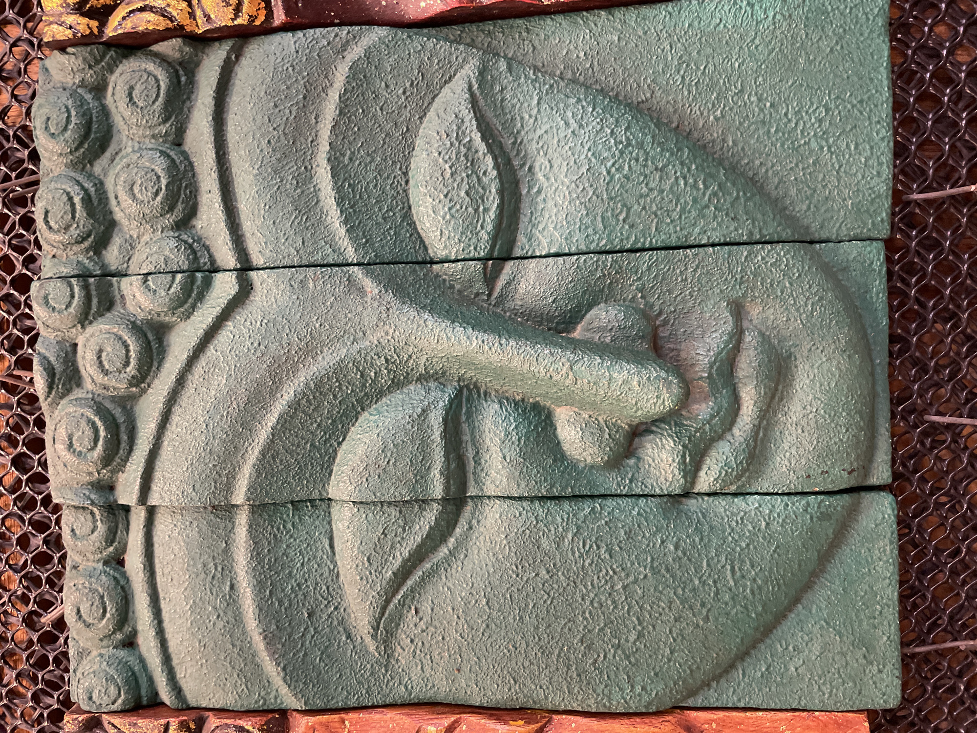 The image displays a close-up, head-on view of a fragmented, teal-colored relief sculpture of a Buddha face. The sculpture is divided into three vertical sections, with visible seams between them. The left and right edges of the sculpture are bordered by roughly textured, reddish-brown and gold-patterned elements. The Buddha's eyes are depicted closed, with subtle lines indicating the eyelids and eyebrows. The nose is prominent, and the mouth is slightly parted. The forehead is adorned with stylized curls, reminiscent of hair. The background consists of a dark, woven mesh. The lighting casts shadows that highlight the textured surface of the sculpture. There are no people present in the image. The sculpture appears to be a decorative item, possibly for sale or display in an indoor setting. Its specific location in Pattaya City, Thailand, is not discernible from the visual information provided.