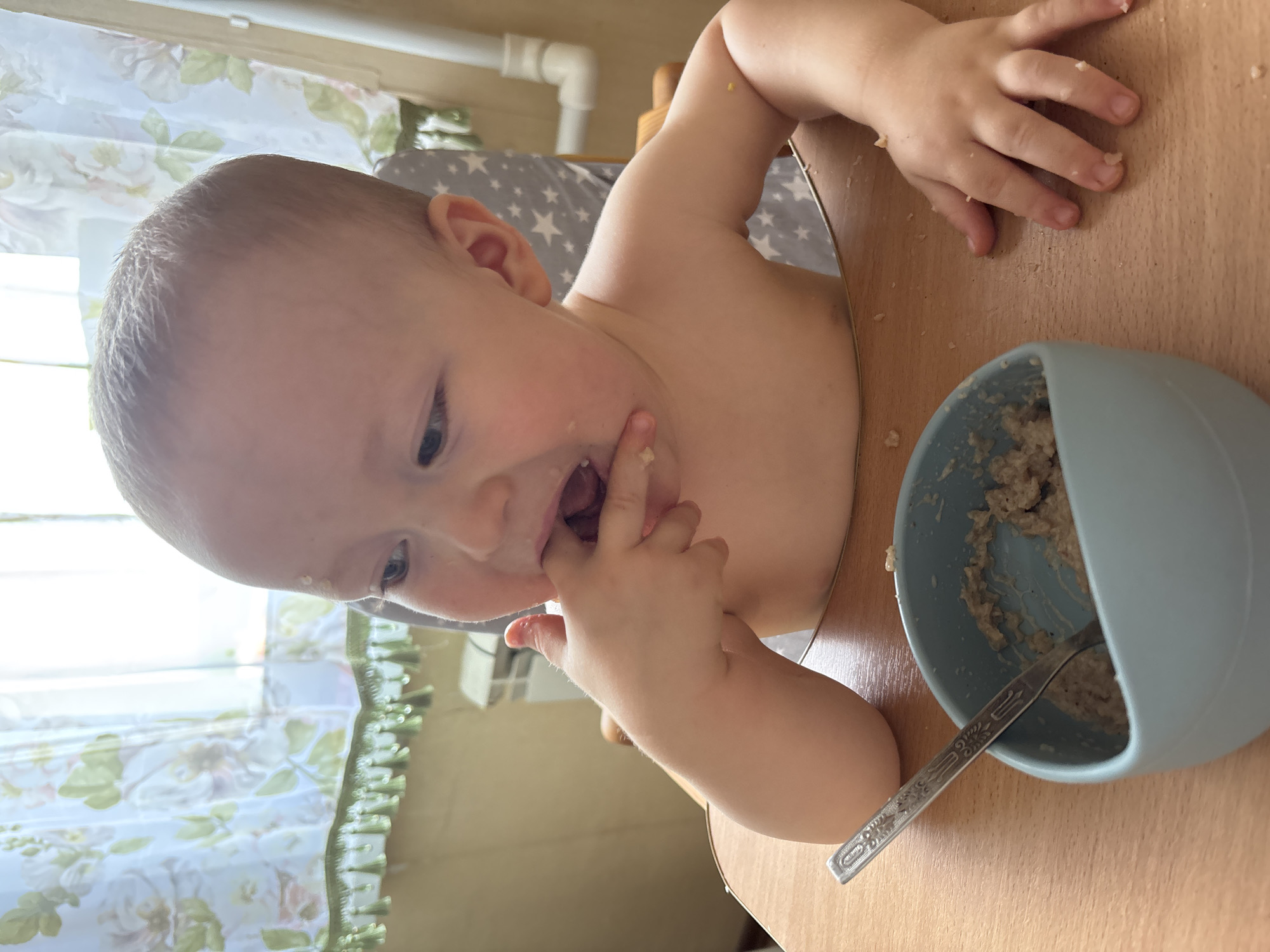 A high-angle, close-up shot shows a baby boy sitting at a wooden table, with a light blue bowl of oatmeal and a spoon in front of him. The baby is wearing a bib and has oatmeal on his face and hands. He is reaching into the bowl with his fingers, as if to feed himself. The background shows a window with floral curtains and a grey and white star-patterned high chair. The lighting suggests it's daytime. The baby appears focused on his food, with a hint of a playful expression. There is no visible text in the image. The context indicates the photo was taken in Naberezhnye Chelny, Russia.