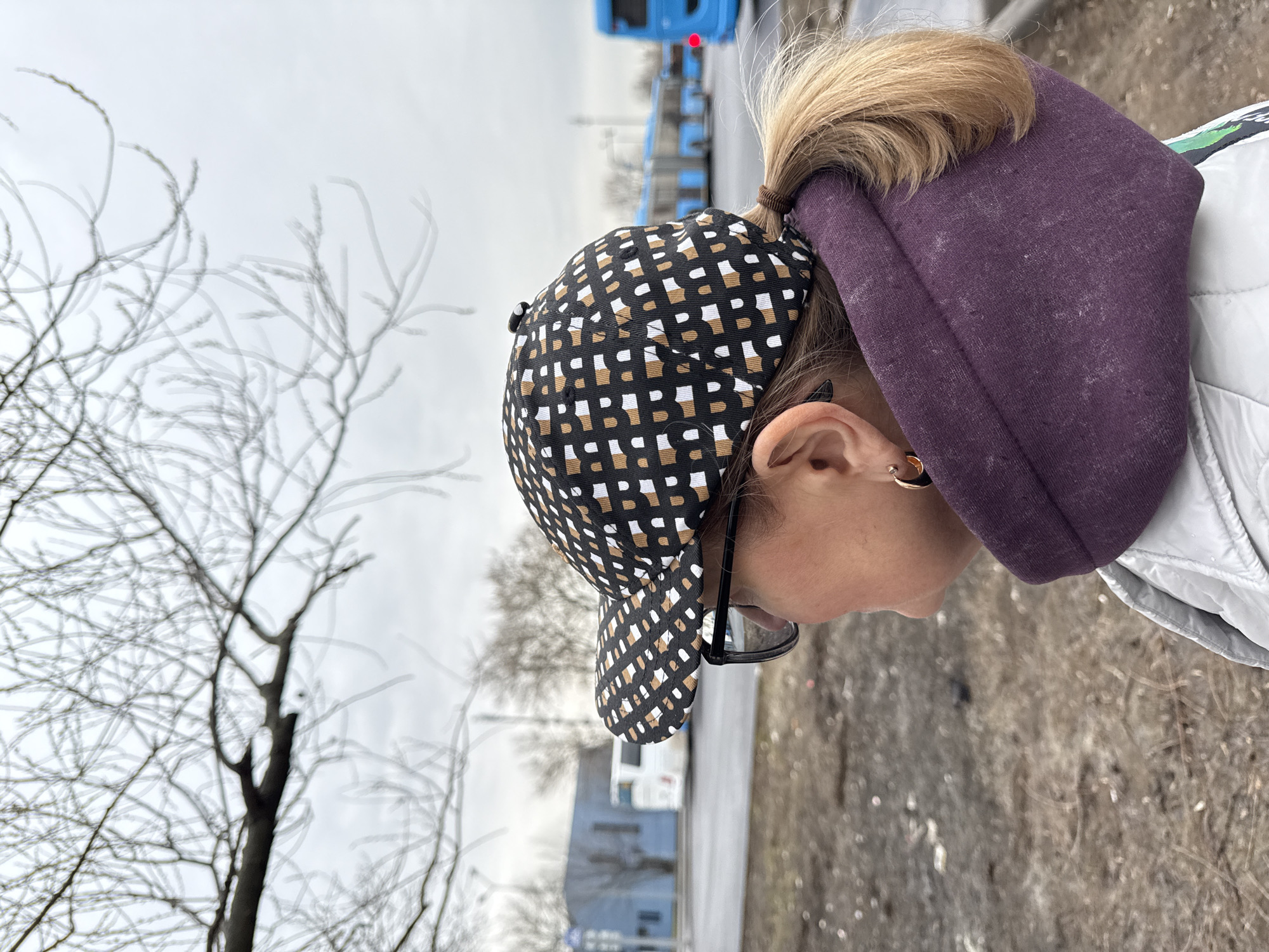 A woman with blonde hair in a ponytail, wearing a patterned baseball cap and a purple hoodie, stands outdoors. She has eyeglasses and an earring in her left ear. To her left, a bare tree is visible against a gray, overcast sky. In the background, a street scene includes blue vehicles and buildings, typical of an urban environment. Based on the visual cues, this location resembles an urban area in Moscow, Russia. The ground appears to be covered in gravel or a similar material.