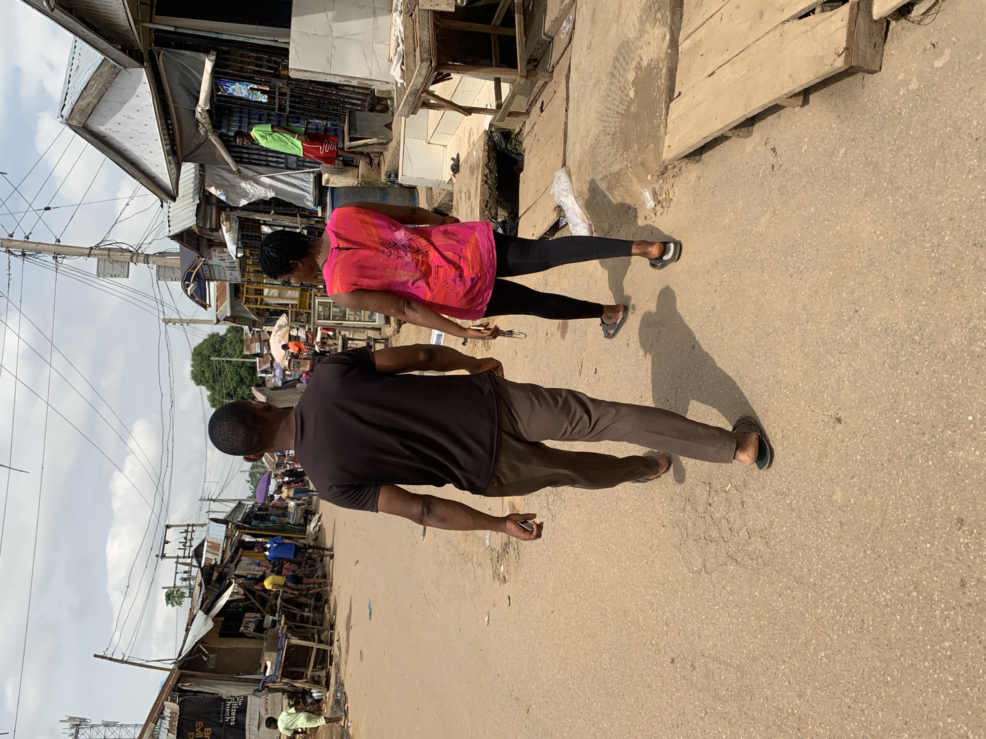 Two people walk along an unpaved road in Abuja, Nigeria. The person in the foreground wears brown pants and a black shirt and carries a mobile phone. The person behind them wears black leggings and a bright pink shirt. Shops and stalls are lined along both sides of the road. Overhead power lines crisscross the sky.