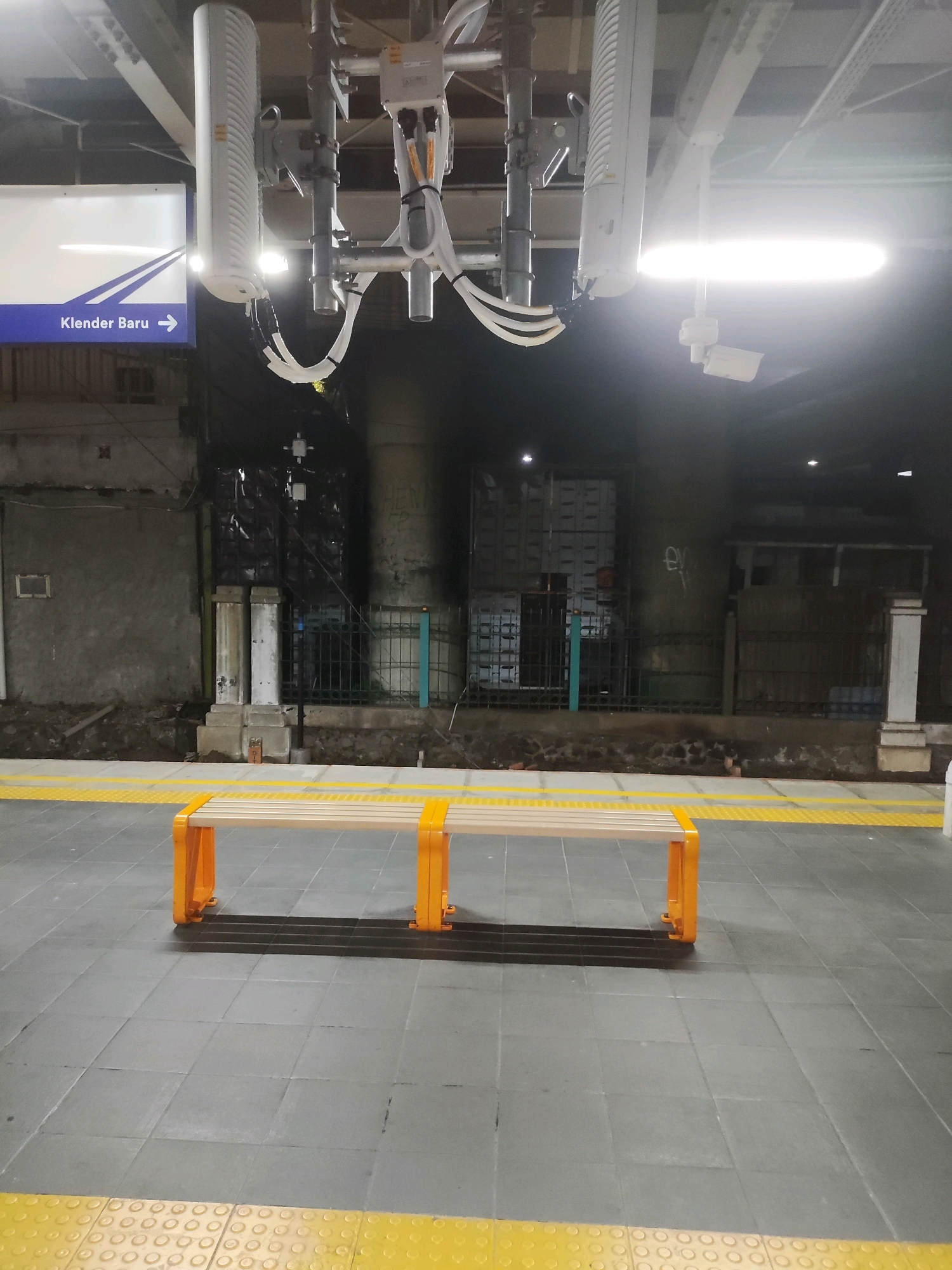 The image depicts a train platform at Klender Baru Station in Bekasi, Indonesia. In the foreground, a modern, orange-legged bench with a light-colored slatted seat is centrally placed on a grey tiled floor. A yellow tactile paving runs along the edge of the platform. Overhead, a structure supports what appear to be communication antennas and lighting fixtures, along with a security camera. The background shows a dark, somewhat grimy wall with visible pipes and conduits, suggesting an industrial or utilitarian area behind the station platform. The lighting is artificial, with a bright strip light illuminating the overhead equipment. There are no people visible in the scene.