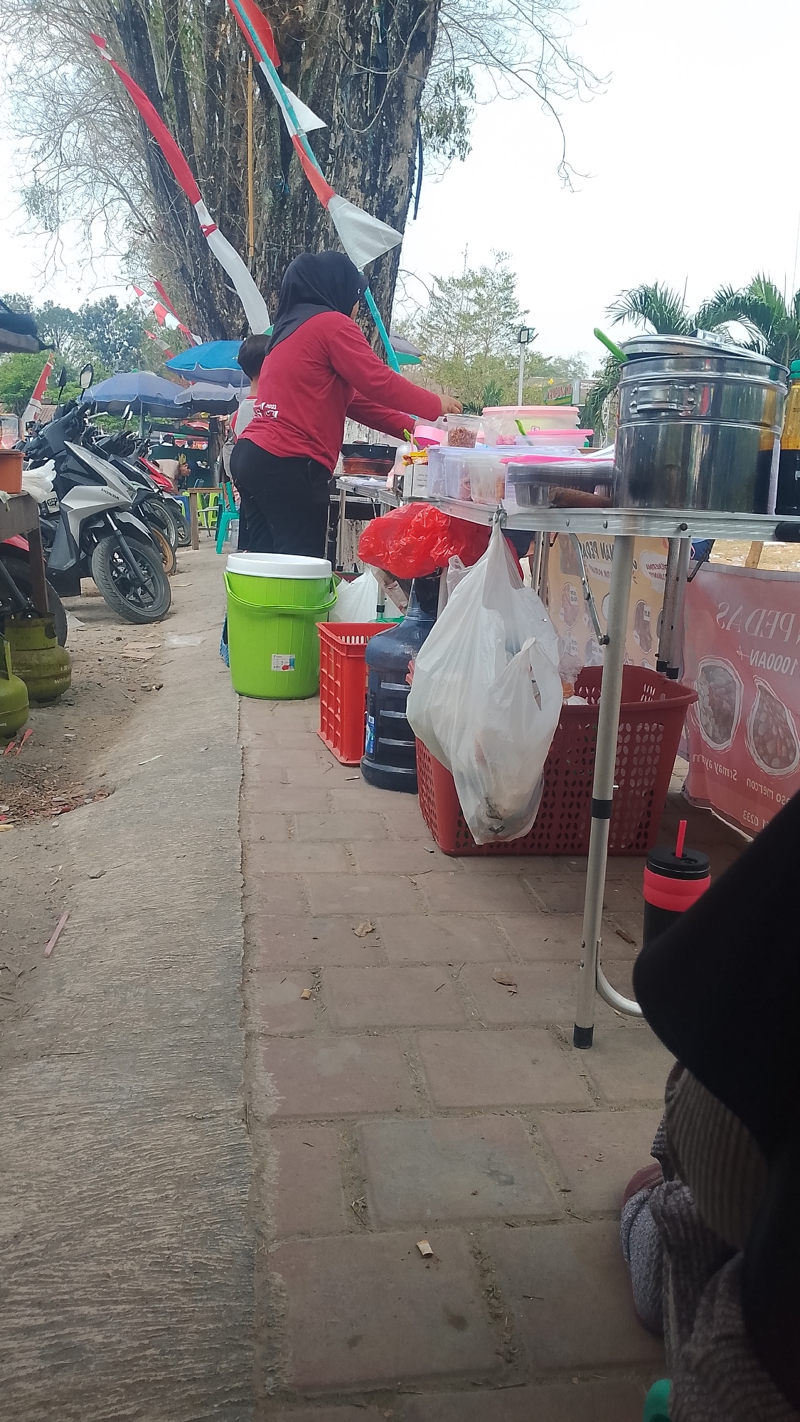 A woman wearing a red long-sleeved shirt and a black hijab is standing at a food stall. She is facing away from the camera and appears to be preparing food. The stall has various containers of food, a large metal pot, and other items for sale. In the background, there are motorcycles, trees, and Indonesian flags. The stall is set up on a paved area next to a sidewalk. The overall scene depicts a street-side food vendor in Karanglangu, Indonesia.