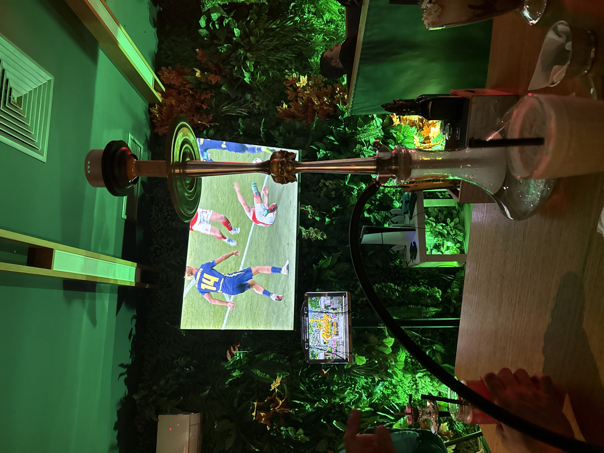 Here is a matter-of-fact description of the image:
The image depicts an indoor scene, possibly a restaurant or lounge, adorned with greenery and faux foliage, creating an immersive, garden-like atmosphere. A hookah is prominently featured, with its base resting near a table where drinks, including what appears to be a milkshake, are placed. A television screen is displaying a sports game, specifically a soccer match. Another smaller screen displays a landscape advertisement of some kind. The environment has a distinct green hue, likely due to the lighting and the abundance of plants, real or artificial. A vent is present on the wall. A hand appears to be holding a part of the hookah pipe.