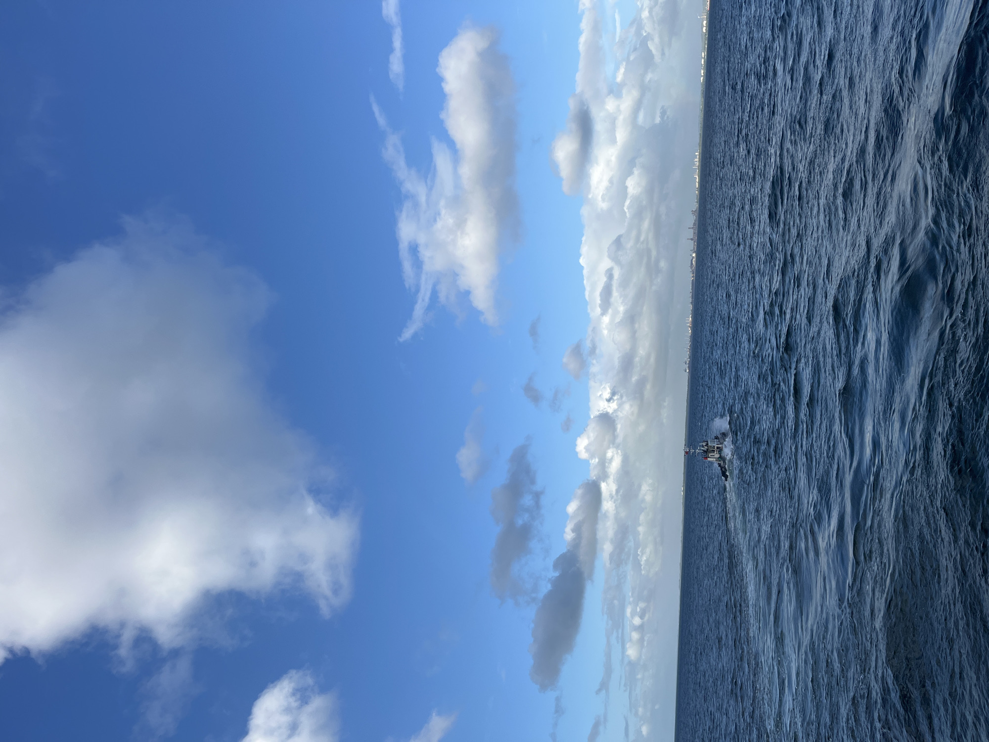 The image shows a seascape with a clear blue sky partially covered by white, puffy clouds to the left, transitioning to a more overcast sky with heavier clouds on the right. The sea is choppy with dark blue, textured water. A ship or boat is visible in the distance, slightly to the right of the image's center. In the background, a faint line of what appears to be a city skyline is visible along the horizon. The location appears to be near the city of Unknown, Unknown.