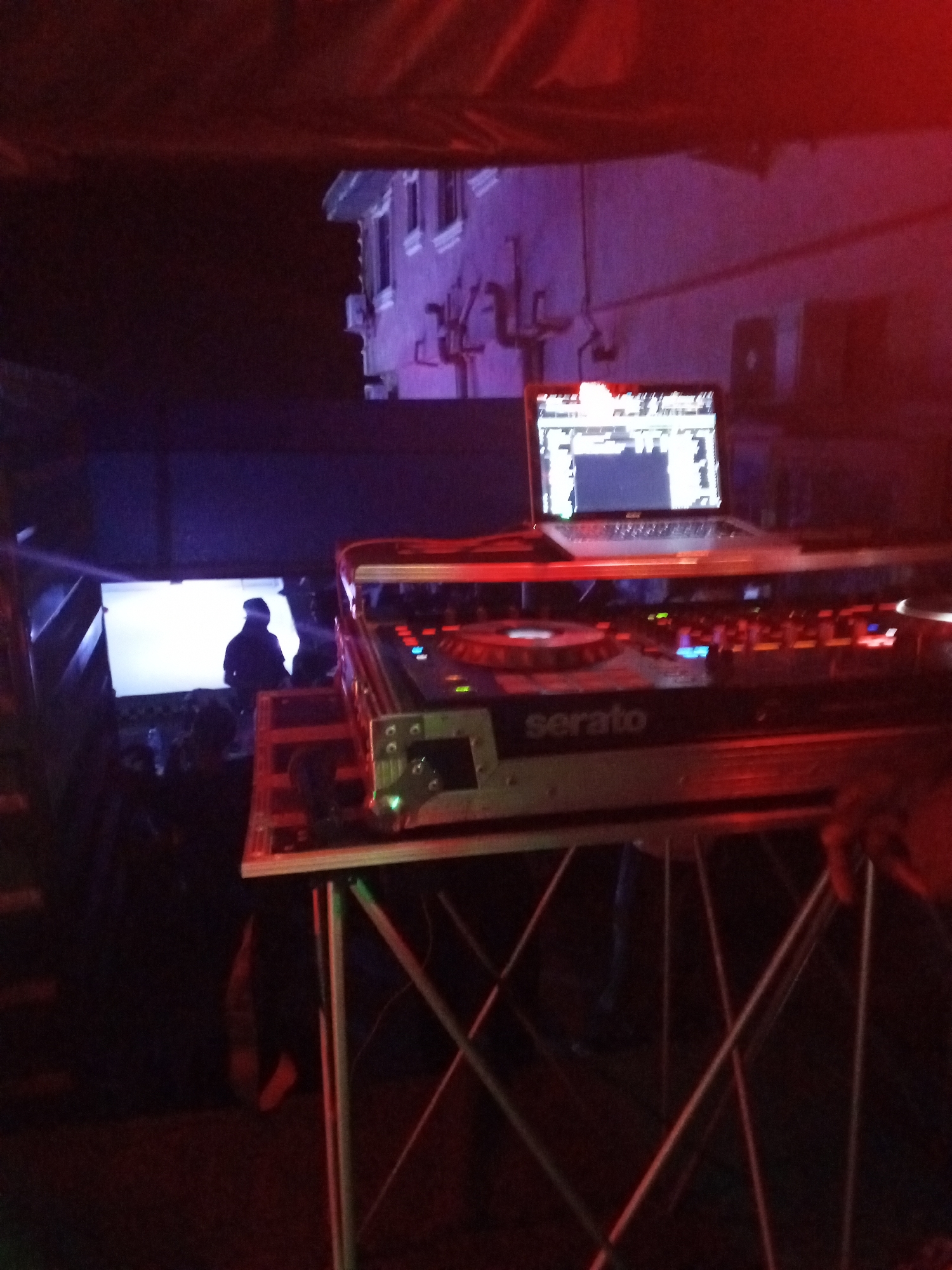 The image shows a dimly lit scene, likely at night, with a DJ setup in the foreground. A laptop with a bright screen is positioned atop a Serato DJ controller on a metal stand. Behind the DJ setup, a person is silhouetted against a bright screen or light source. In the background, a multi-story building is visible, possibly a residential structure, suggesting an urban environment. The red and purple lighting suggests a club or event setting. The location is Lokoja, Nigeria, based on the prompt. Specific details about interactions or actions are not clear.