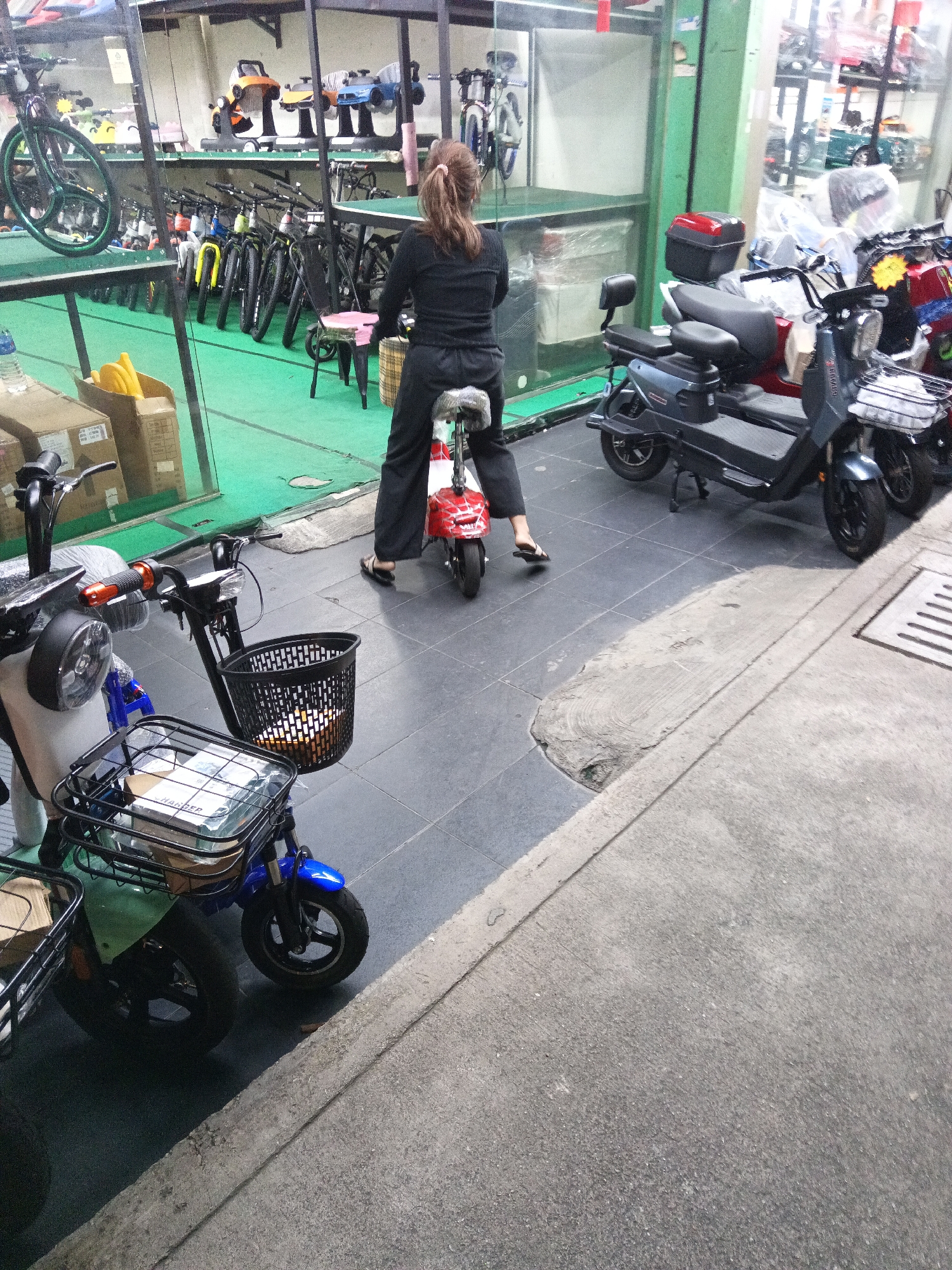 A woman with brown hair pulled back is sitting on a small red electric scooter in a shop. She is wearing a black long-sleeved shirt and black pants. She is facing away from the camera, looking towards a display of bicycles and other electric scooters. The shop appears to be an outdoor market or a sidewalk store in Kuala Lumpur, Malaysia, with various items for sale. The ground is tiled with gray squares and there is a concrete sidewalk in the foreground. The scene is well-lit, with natural light coming from the open storefront.