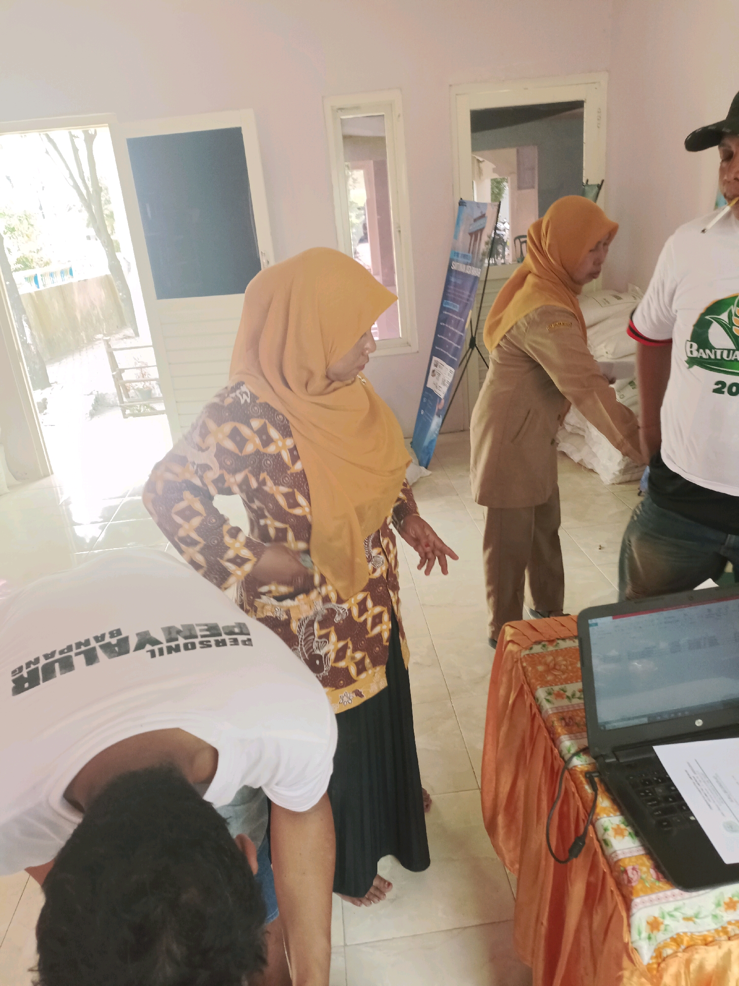 In Guminingrejo, Indonesia, a group of people are gathered indoors. A woman in an orange hijab and a patterned brown and white dress stands in the center, her hand extended as if gesturing. To her left, a person wearing a white t-shirt with black text that reads "PERSONIL PENYALUR BANPAN" is bending over, their back to the camera. To the right, a man in a white t-shirt with the word "BANTUAN" visible and a black baseball cap is smoking a cigarette. Behind the woman in the orange hijab, another woman in a tan uniform with an orange hijab is visible. A table covered with an orange tablecloth and floral accents is in the foreground to the right, displaying a laptop and some papers. The room has light-colored walls and a tiled floor. Natural light streams in from an open doorway or window on the left, illuminating the scene.