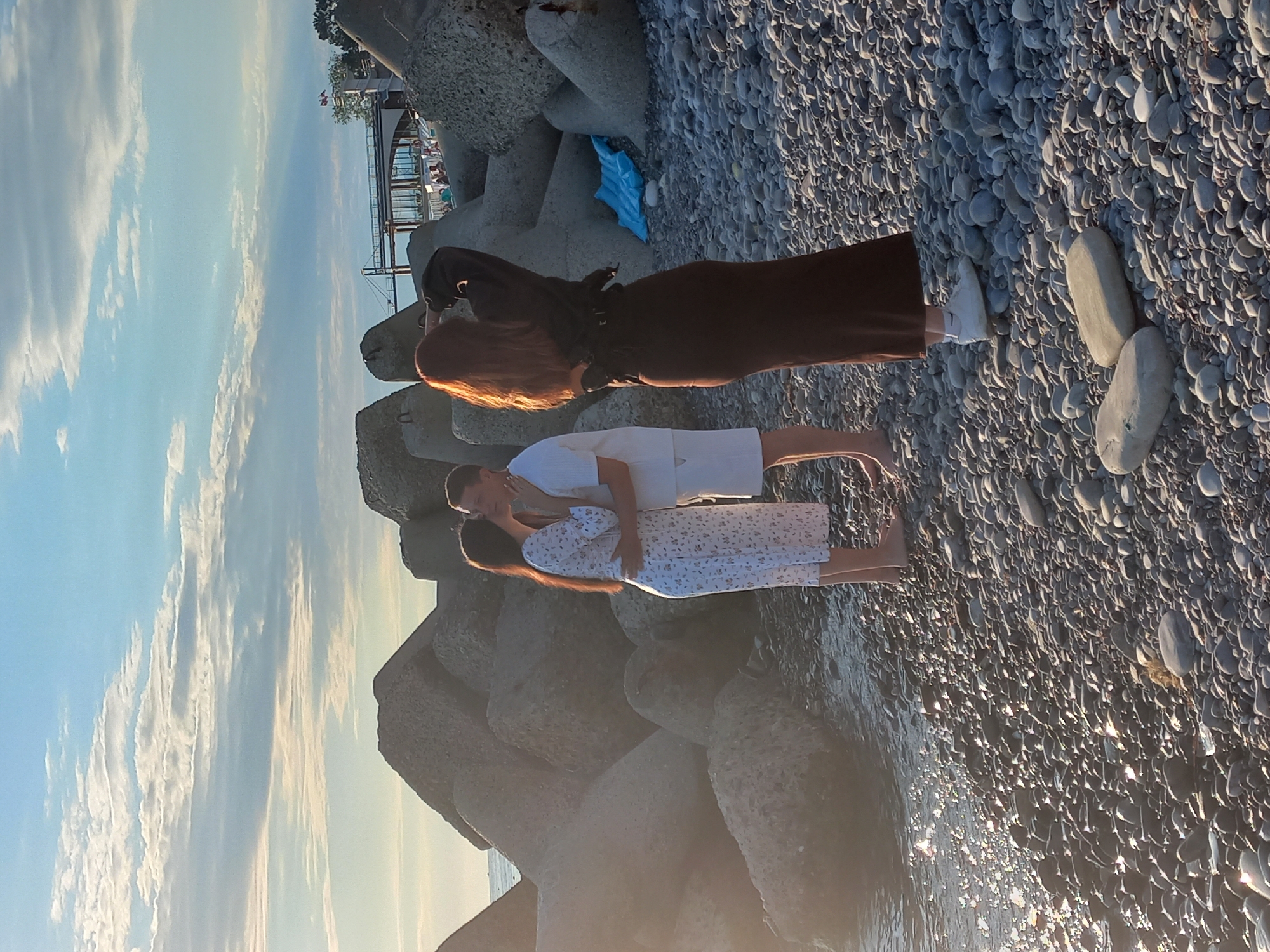A medium shot captures a romantic moment between a man and a woman on a pebble beach in Sochi, Russia, with a third woman observing. The couple, positioned in the center-left of the frame, are embracing and kissing. The woman is wearing a white floral dress and the man is in a white t-shirt and shorts. To the right, a woman with red hair, seen from behind, is standing and looking towards the sea, wearing a brown dress and white sneakers. She has her arm raised, appearing to shield her eyes from the sun. The background features large, angular concrete breakwater structures typical of Sochi's coastline, leading to a distant view of a bridge and buildings under a cloudy sky. The water's surface sparkles with sunlight. The overall scene is set during what appears to be late afternoon or early evening.
