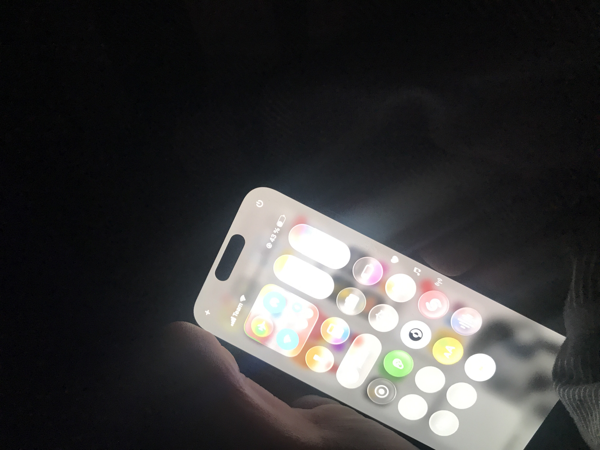 The image shows a person holding a smartphone, likely in a dark environment. The phone screen is illuminated, displaying what appears to be a custom interface or a set of app icons arranged in an unconventional layout. The interface features translucent elements and bright, circular icons. The battery percentage is visible in the upper right corner. Some details like "Team" can be seen in the upper left corner. The person's hand and a sleeve of clothing are visible. There is no information to suggest that this image was taken in Yerevan, Armenia.