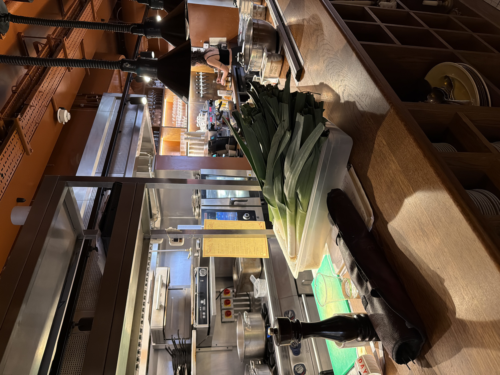 The image depicts the interior of a professional kitchen in London, United Kingdom. In the foreground, a wooden countertop displays a collection of leeks in a clear plastic container, a dark leather knife roll, a clear plastic cup, and a black pepper grinder. Behind these items, stainless steel kitchen equipment is visible, including pots on a stove, ovens, and shelving units stocked with various kitchen supplies. A woman is partially visible in the background, working at a coffee machine. The overall environment suggests a busy, operational restaurant kitchen.