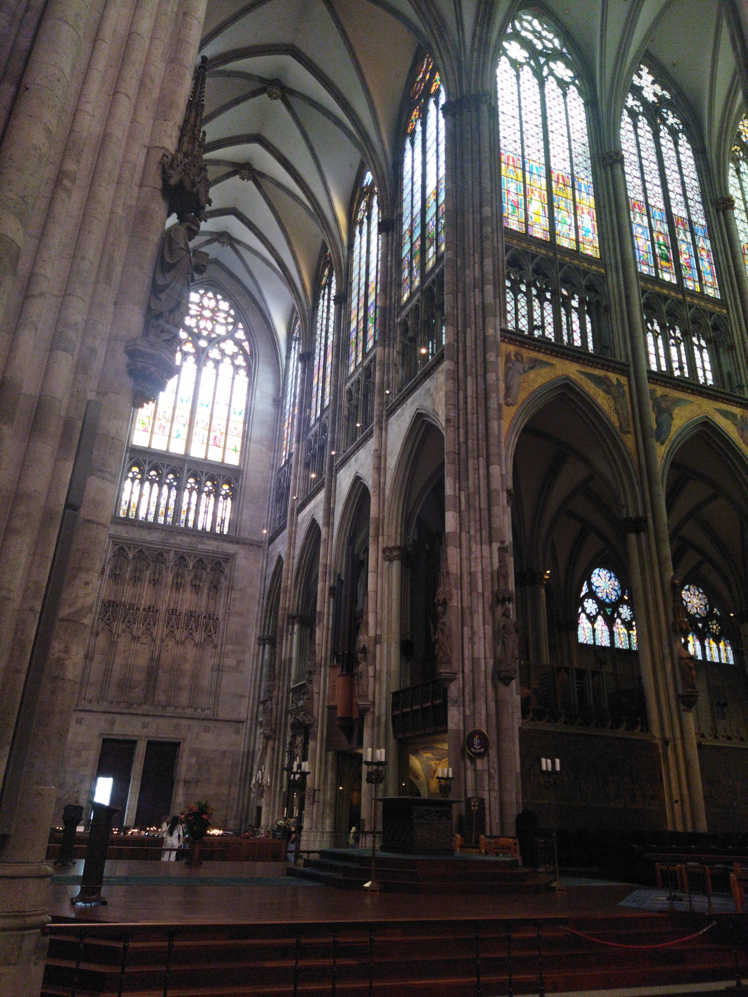The image depicts the interior of a grand cathedral, likely Cologne Cathedral in Germany, given the architectural style. The perspective is from the lower nave, looking upwards and towards the apse. The scene is characterized by soaring Gothic architecture, with massive stone pillars supporting ribbed vaults and intricate arched windows filled with stained glass. Sunlight streams through the stained glass, casting colorful patterns on the stone surfaces.
In the foreground, wooden pews are visible, arranged in rows on a wooden floor. Towards the middle ground, an altar area is set up, with a raised platform and various religious artifacts. Several people are present in the altar area, some appearing to be in clerical attire, possibly engaged in a service or ceremony. One individual in a white garment stands near a floral arrangement. Candles are lit on stands around the altar, adding to the ambiance.
The walls are adorned with ornate carvings and decorative elements, typical of a historic cathedral. The scale of the building is immense, emphasizing its architectural grandeur and religious significance. The lighting is a mix of natural light from the windows and artificial light from unseen sources, creating a dramatic interplay of light and shadow. The overall impression is one of solemnity, beauty, and historical weight.
The location within the city of Monheim am Rhein, Germany, cannot be definitively confirmed from the image alone, as the architecture is characteristic of many large Gothic cathedrals across Europe. However, if this specific cathedral is indeed in Monheim am Rhein, it would represent a significant historical and architectural landmark within that city.