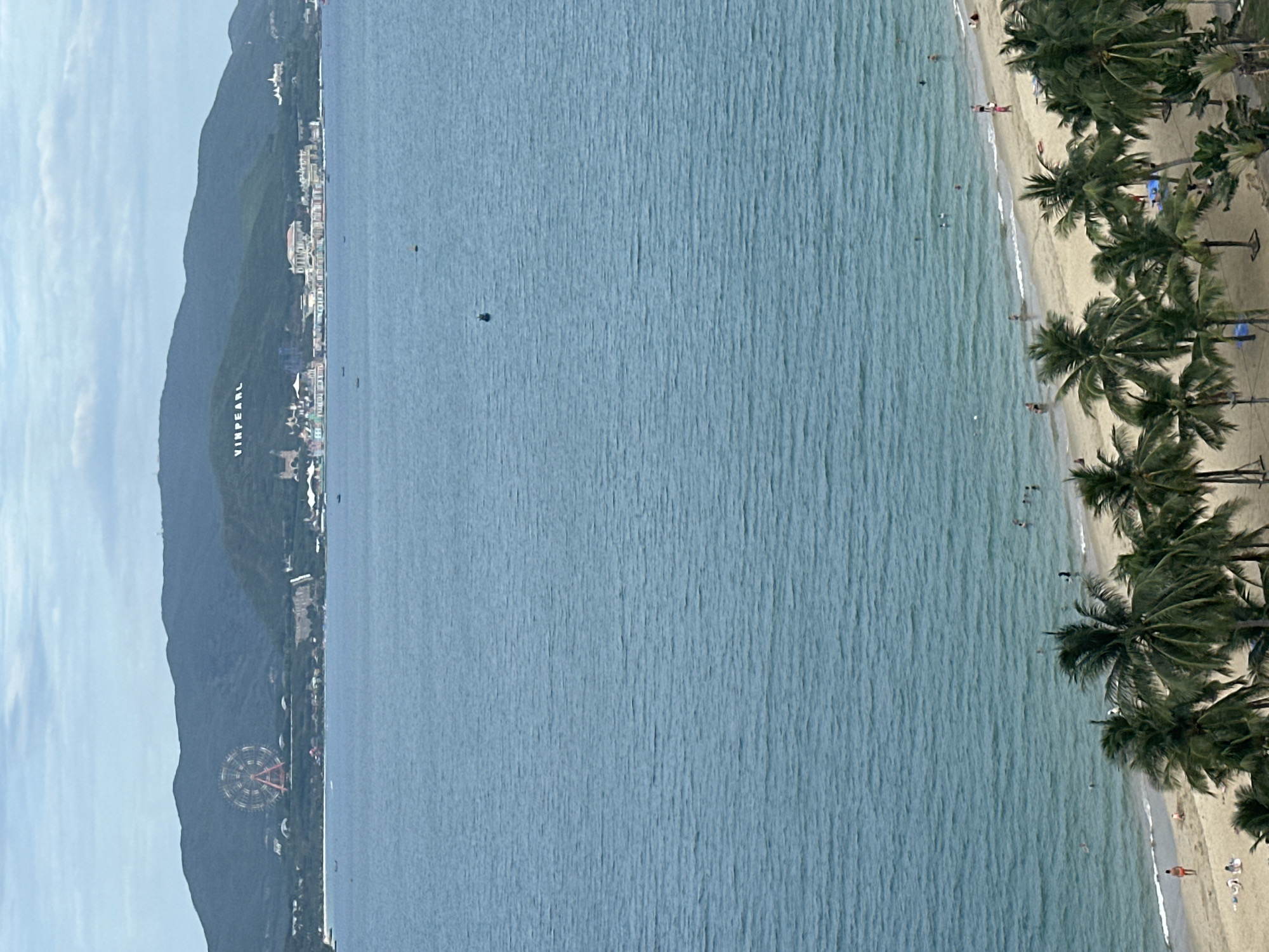 This high-angle, panoramic image captures a vibrant coastal scene in Nha Trang, Vietnam. In the foreground, a sandy beach is visible, densely lined with numerous tall palm trees. Several people are scattered along the shore, some walking, others wading or swimming in the calm, blue-green ocean that meets the beach. The water exhibits gentle ripples, and a couple of small, dark objects, possibly boats or buoys, are present further out.
In the midground and background, a range of dark green mountains dominates the horizon, under a partly cloudy sky. Along the base of these mountains, facing the sea, various buildings and structures are visible. A prominent white sign spelling "VINPEARL" in large block letters is etched onto the side of one of the mountains. Further to the left, a large Ferris wheel with red-orange spokes can be seen nestled against the mountainous terrain, characteristic of the VinWonders resort complex.
The scene depicts a sunny daytime setting, suggesting good weather for beach activities and showcasing a popular resort area. The visible text "VINPEARL" and the Ferris wheel are strong geographical cues confirming the location within Nha Trang. The overall atmosphere appears leisurely and inviting.