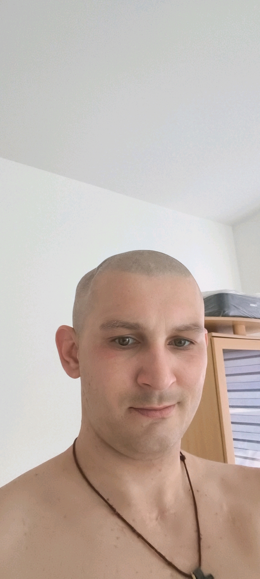 The image shows a close-up of a person with a shaved head. The person is looking at the camera, and their expression is neutral. They are wearing a thin brown cord necklace. The background is a plain white wall, with a wooden cabinet and a black object on top of it visible on the right side of the image. The lighting is bright and appears to be natural. The scene is an indoor setting, possibly a residential room. The specific location in Wolgast, Germany, cannot be determined from the image alone.