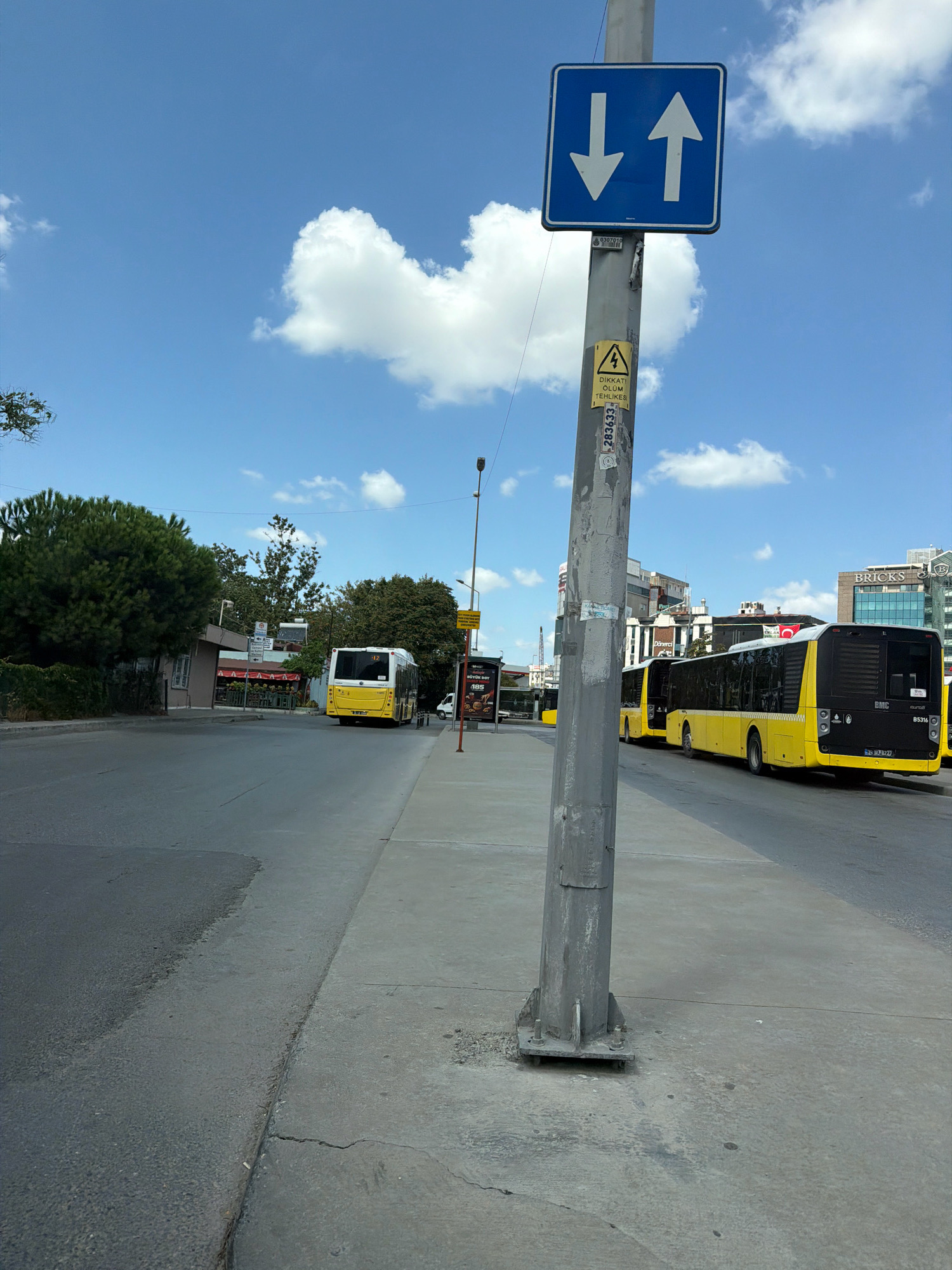 The image depicts a street scene in Bakırköy, Turkey, on a clear day with scattered white clouds. A grey metal utility pole stands prominently in the foreground, supporting a blue traffic sign with two white arrows, one pointing up and one pointing down, indicating a two-way traffic street. Attached to the pole are several stickers, including a yellow triangular warning sign with a lightning bolt symbol and Turkish text.
The street is paved and appears to be a bus route, with yellow public buses visible in the background. One bus, a BMC model with license plate 34 JY 2777, is parked along the side of the road, while another is further down the street, also yellow and white. Buildings line the street, with some appearing to be shops or businesses, and others are taller structures in the distance. Trees with green foliage are present on the left side of the image. The sidewalk is made of concrete and shows signs of wear and tear, with cracks and some loose gravel. The overall impression is of an urban environment during daylight hours.