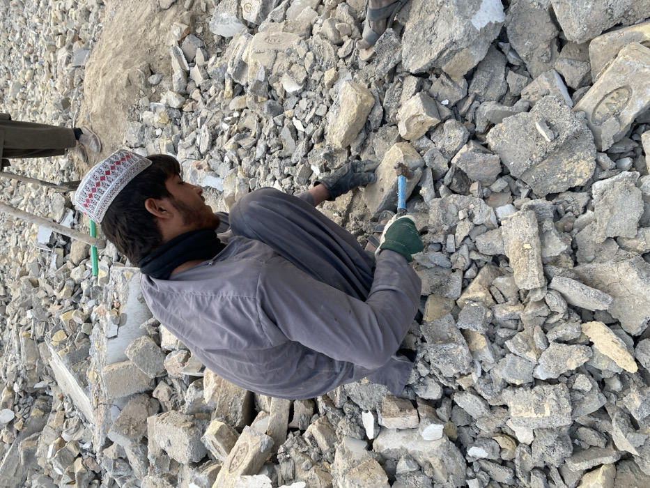 The image depicts a debris-filled site in Kandahar, Afghanistan, consistent with a demolition or post-destruction environment. A young male, appearing to be in his late teens or early twenties, is centrally positioned and crouched low. He wears a grey long-sleeved tunic and trousers, a black neck gaiter, and a white skullcap adorned with red geometric embroidery. His left hand, gloved in green, holds a blue-handled tool, resembling a hammer or chisel, which he is actively using to strike a light-colored brick fragment situated among a dense pile of grey and beige concrete rubble and broken bricks. His right hand, covered by a dark glove, is also interacting with the debris. In the background, partially visible above the main subject's shoulder, are the lower legs and feet of a second individual, wearing dark trousers and light-colored shoes, standing on a small mound of dirt and holding a long tool handle, likely from a shovel or pickaxe. The entire visible area is covered in various sizes of construction debris.