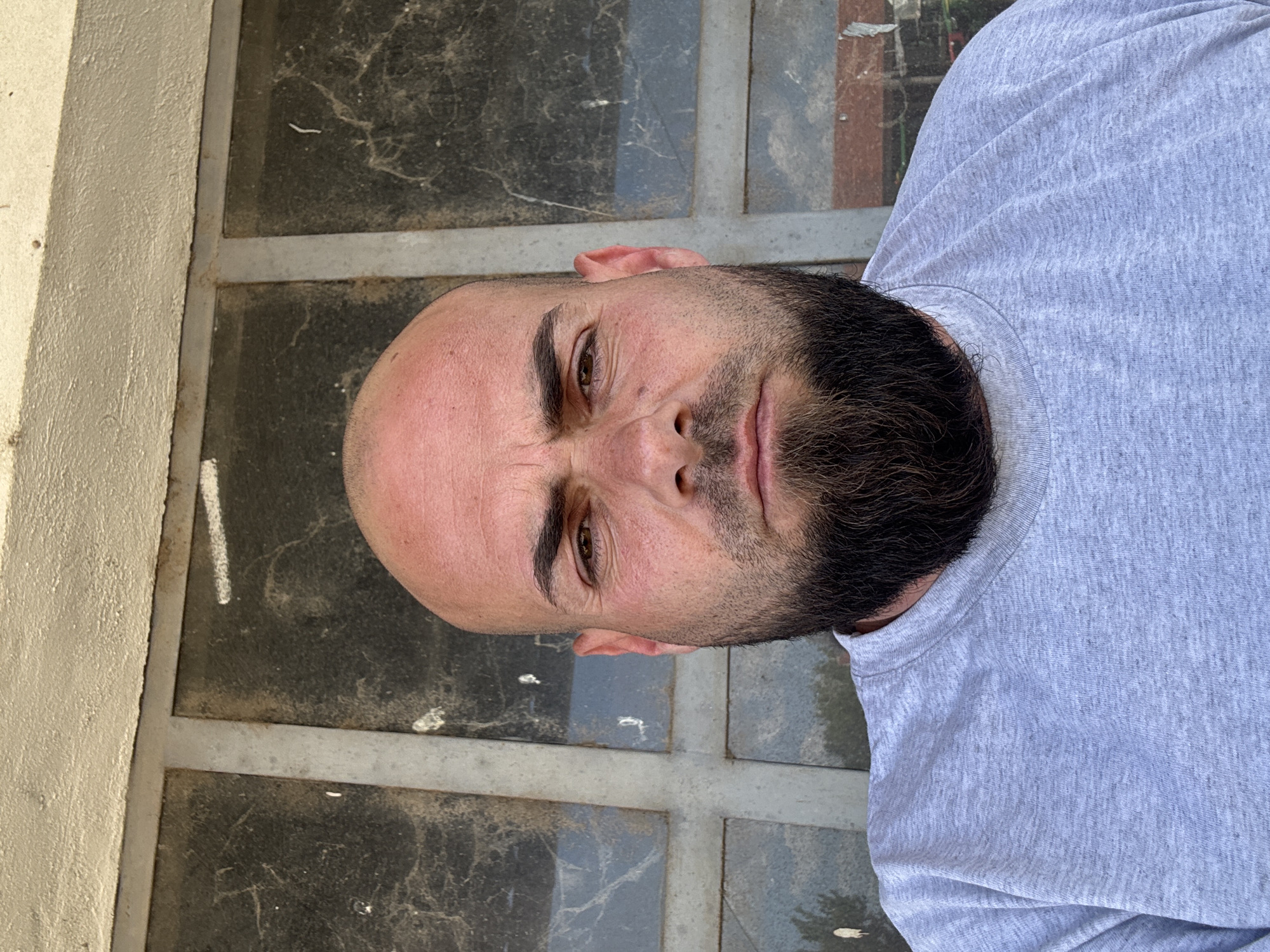 A middle-aged man with a bald head and a full, dark beard is looking directly at the camera. He appears to be standing outdoors, near a weathered, multi-paned window. The man is wearing a grey crew-neck t-shirt. The location is possibly Sarnevo, Bulgaria.