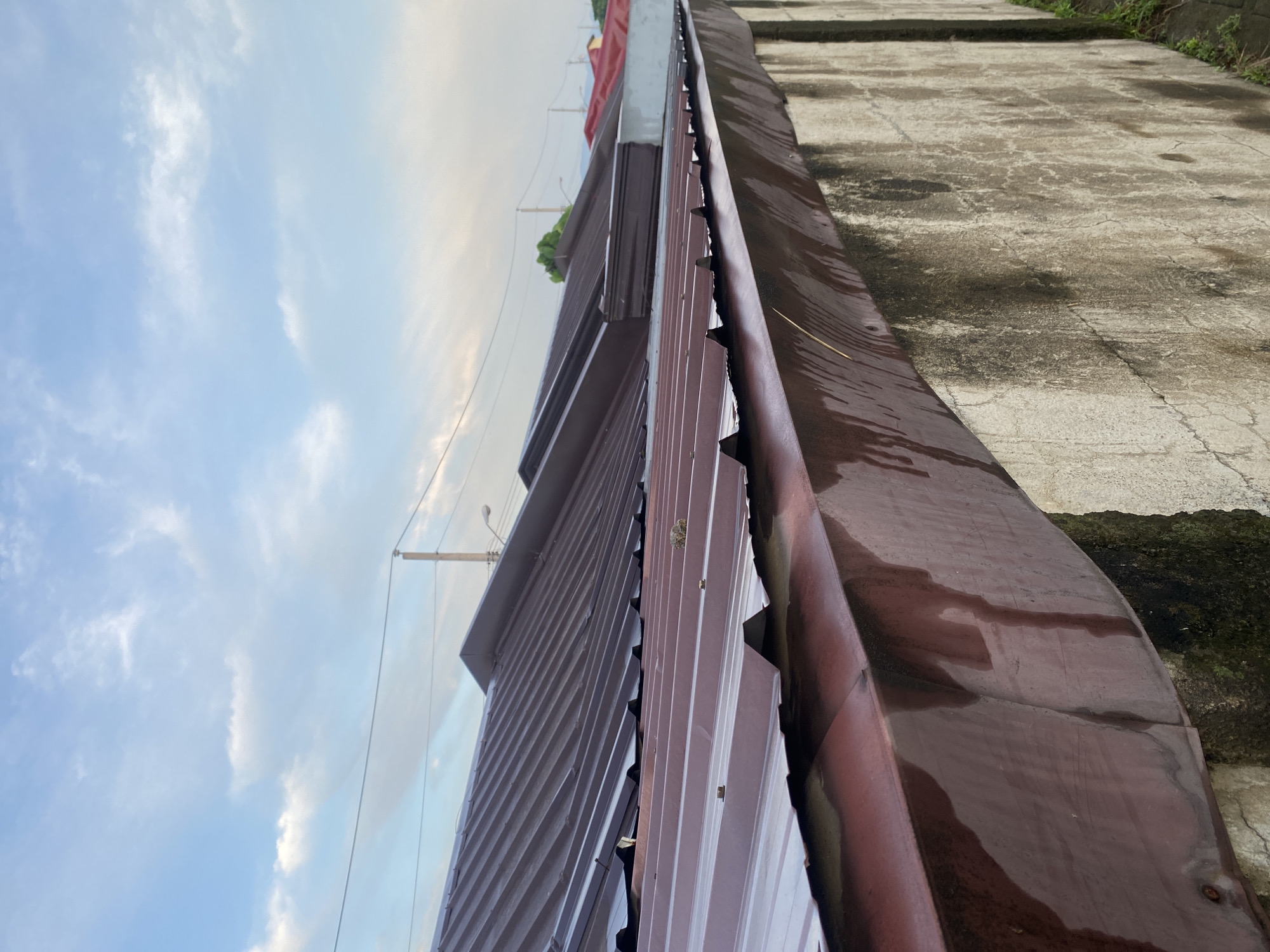 The image shows a view of a building with a corrugated metal roof. The roof appears to be two-toned, with one section a dark brown and another a lighter gray or silver color. A utility pole and wires are visible against a cloudy blue sky. The image is taken from a low angle, looking up at the building.
The location is inferred to be Davao City, Philippines, based on the request. However, the image itself doesn't contain any specific elements confirming this location.